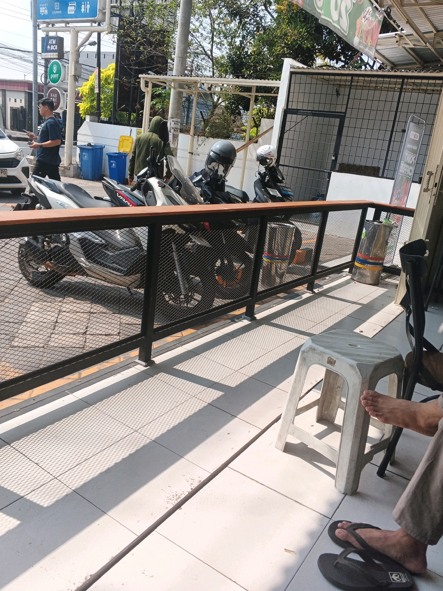 In Genuk Barat, Indonesia, a scene unfolds with motorcycles parked in front of a building. A person in a green hoodie stands near the motorcycles, while another individual in a dark blue shirt and shorts is walking away from the viewer, holding a phone. A white plastic stool is placed on the tiled ground, with a person's feet and flip-flops visible near it. Sunlight casts shadows across the scene, highlighting the textures of the pavement and the metal railing. Trash cans and signage are also present in the background, indicating a public or commercial area. The overall atmosphere suggests a casual, daytime setting on a street in Indonesia.