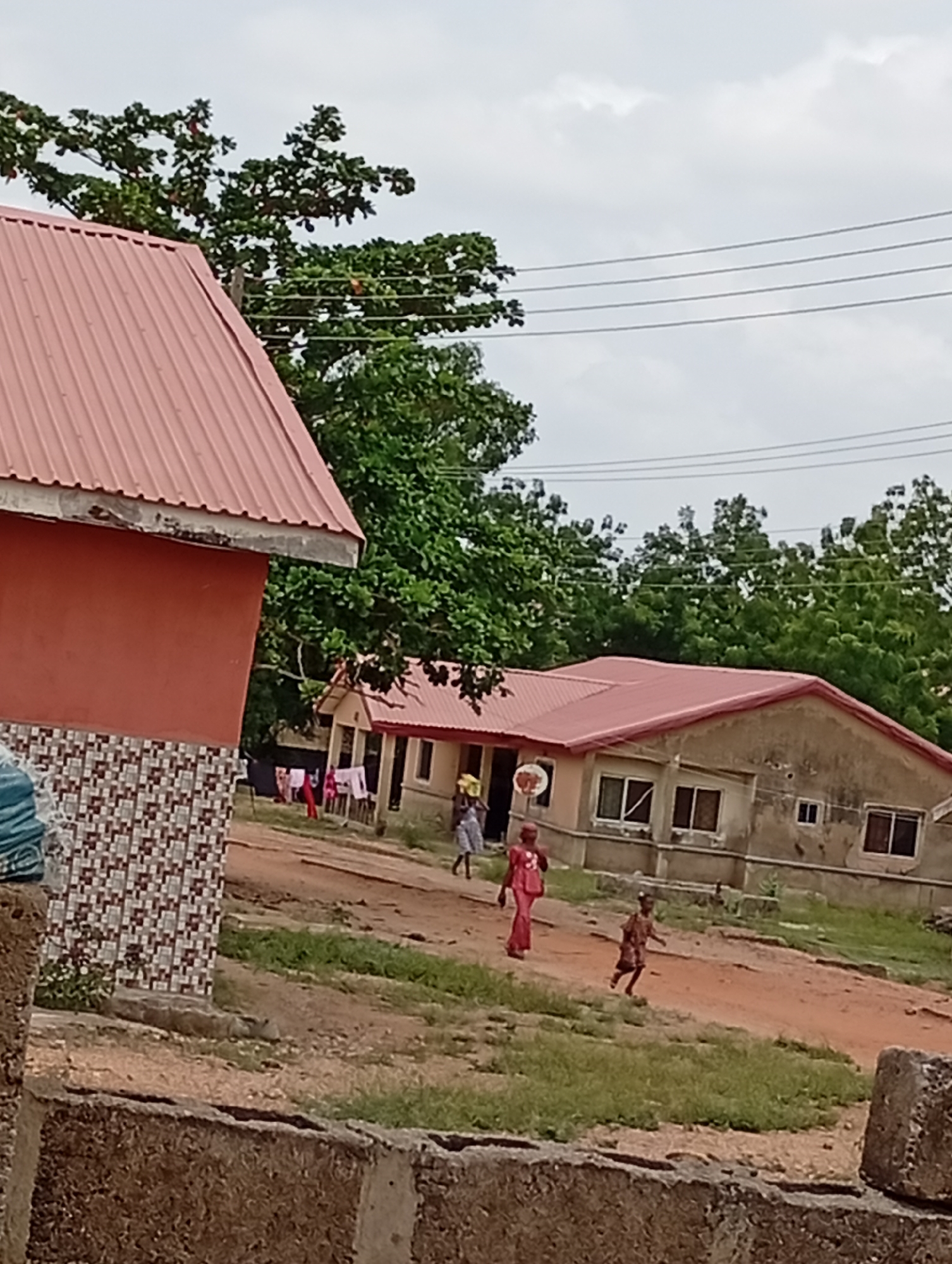 The image shows a residential scene in what appears to be Adankolo, Nigeria. In the foreground, there is a partially visible building with a red roof and patterned tiles. A low concrete wall or foundation is visible at the very bottom of the frame.
In the middle ground, a dirt road or path is visible. Three people are present: one in a red outfit walking with something on their head, another carrying something on their head, and a child running. There are several buildings with red roofs situated along the path. Laundry is hanging outside of one of the buildings.
The background features green trees and a cloudy sky. Overhead power lines are also visible.