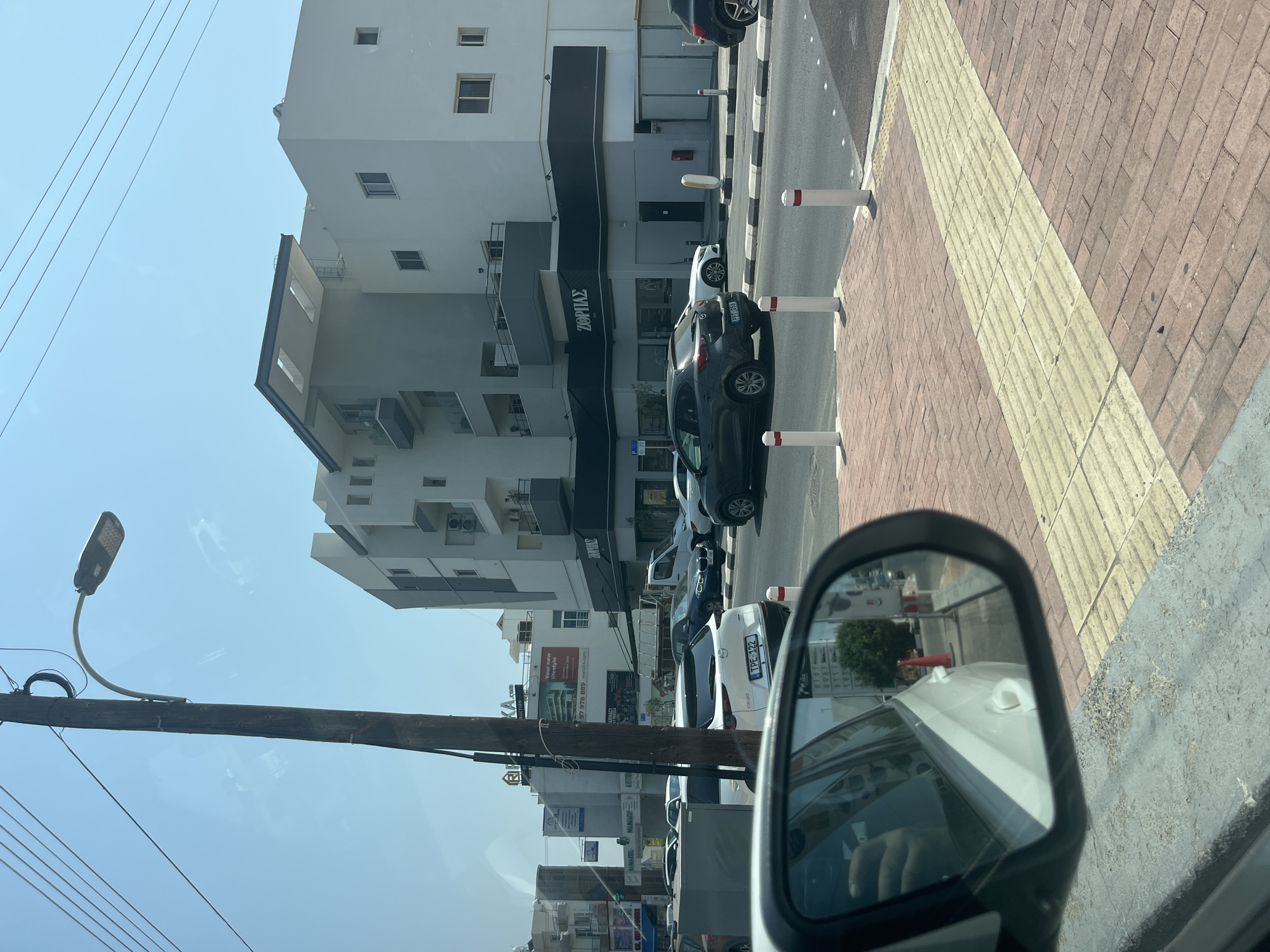 The image depicts a street scene in Germasogeia Municipality, Cyprus, on a clear, sunny day. In the foreground, the side mirror of a white car reflects a portion of the street and sidewalk. The mirror shows the reflection of the car's interior, including a hand on the steering wheel. A utility pole with a street light stands prominently on the left side of the image.
In the mid-ground, traffic moves along a multi-lane road. Several cars are visible, including a white Mazda CX-5 with license plate TPE 122, a dark grey Mazda, and a white BMW. Buildings line the street, featuring modern architecture with balconies and large windows. One building displays a sign that reads "ΖΩΡΠΙΑΣ" in Greek, indicating a commercial establishment. The street is paved with asphalt, and sidewalks are made of brick and tactile paving.
The background shows more buildings and a clear blue sky. The overall impression is that of a typical urban environment in Cyprus, with active traffic and commercial activity.