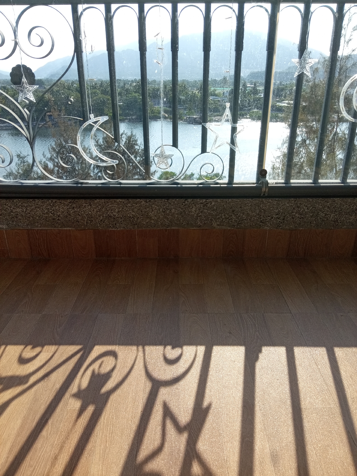 The photograph depicts a scene from a balcony in Nha Trang, Vietnam. The foreground features a tiled floor with prominent shadows cast by a wrought iron railing, which forms the midground. Through the railing, the background reveals a body of water, likely a river or bay, bordered by lush vegetation and glimpses of buildings. Distant mountains are visible on the horizon. Decorative star and moon-shaped ornaments are suspended from the railing, adding a touch of festive atmosphere. The scene suggests a sunny day, with strong shadows and a bright outdoor light.