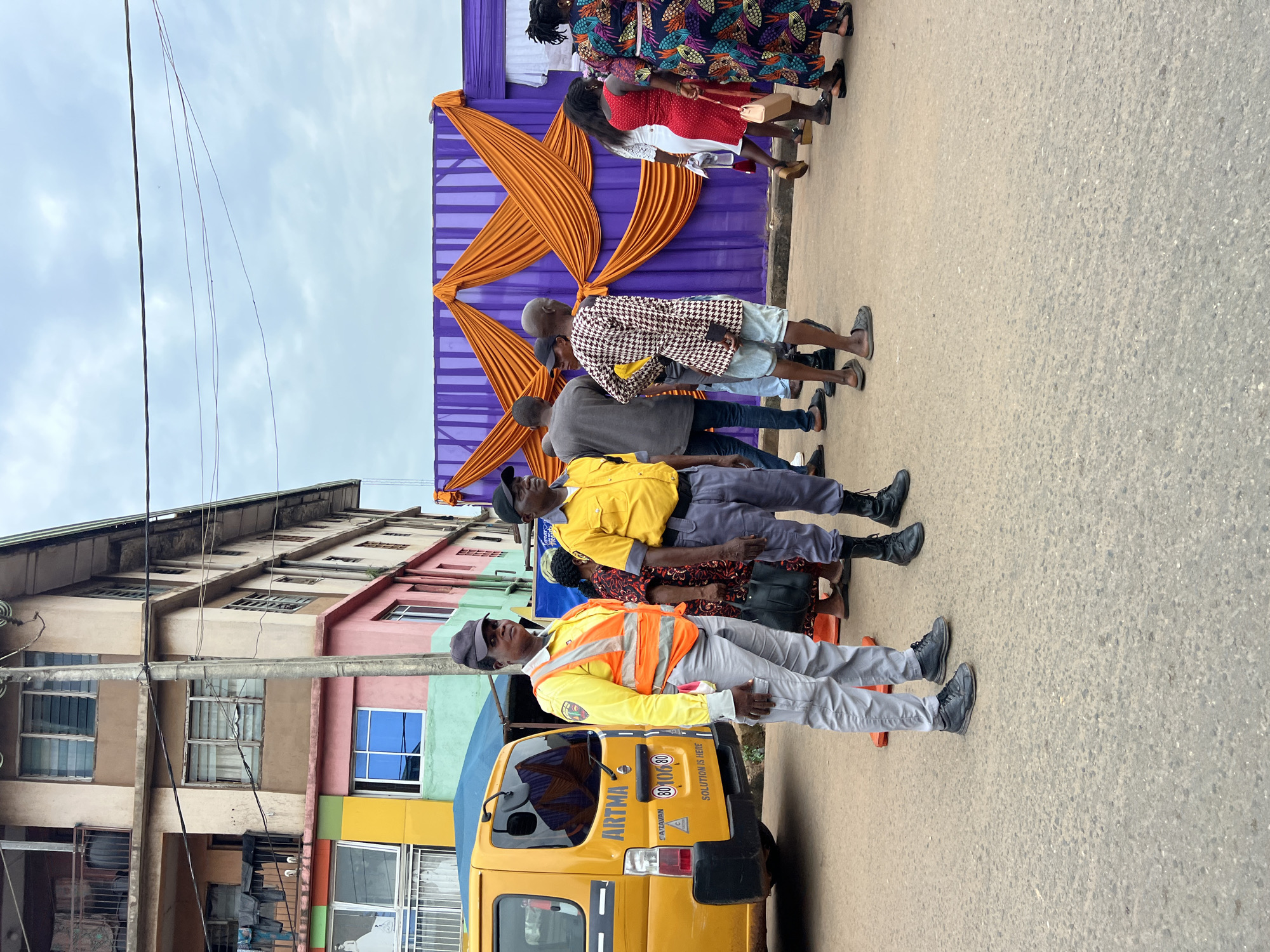 The image depicts a street scene in Nkpor, Nigeria, likely near a venue decorated for an event. Two individuals in what appear to be security uniforms, one woman and one man, stand prominently in the foreground. The woman is wearing a yellow long-sleeved shirt, a reflective orange vest, grey trousers, and a dark cap, with dark shoes. The man beside her wears a yellow collared shirt, grey trousers, and black boots, also with a dark cap. Behind them, a yellow van with "ARTMA" and "CARAVAN SOLUTION IS HERE" visible on its rear, is parked.
A crowd of people are seen in the middle ground, some walking, others standing. A notable feature is a large purple fabric backdrop decorated with draped orange fabric, suggesting an event setup. The background shows a multi-story building with varying colors and balconies, typical of urban architecture in Nigeria. The sky is overcast with a few thin clouds. The ground is asphalt, indicating a road or paved public space. Several traffic cones are also visible near the security personnel.