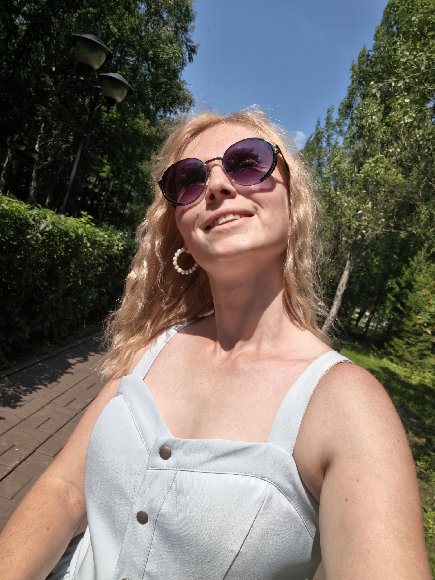 The image shows a woman with blonde, wavy hair wearing sunglasses and a light blue dress. She is standing on a wooden walkway in a park-like setting with trees and greenery in the background. The sky is clear and blue. There are two lamps on a pole visible in the upper left portion of the image. The woman is smiling and looking up. The lighting suggests it is daytime.