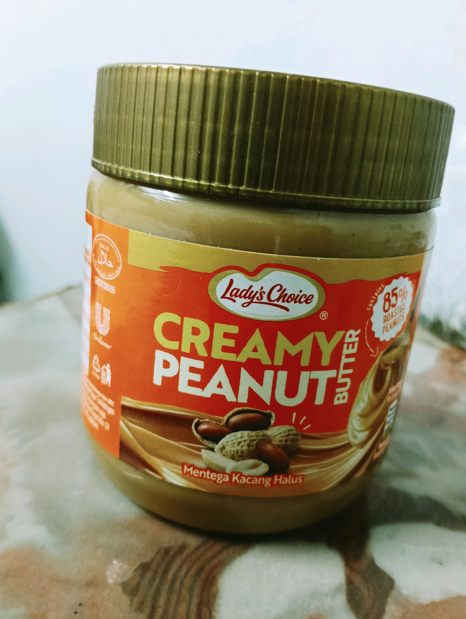 A jar of Lady's Choice Creamy Peanut Butter is presented in a close-up, slightly angled shot. The jar has a golden ridged lid and an orange label with yellow accents. Prominently displayed on the label are the brand name "Lady's Choice," the product name "Creamy Peanut Butter," and the text "85% ROASTED PEANUTS" within a circular emblem. An illustration of peanuts and swirling peanut butter is visible on the label. The jar sits on a patterned surface, possibly a table, with a blurred background. The lighting is soft, creating subtle shadows. There are no people or actions depicted in the image. The image is a product shot and does not provide any information about its specific location within Kuala Lumpur, Malaysia.