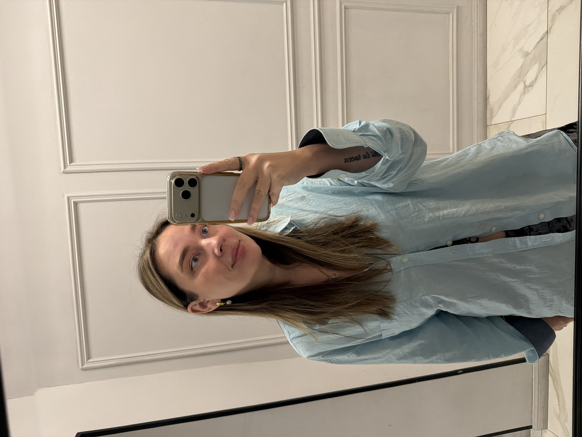 A woman stands indoors, taking a selfie in a mirror. The background includes a wall with decorative panels and a section with marble-like texture. The woman is wearing a light blue button-down shirt. She is holding a smartphone with a triple-lens camera, taking a photo of herself. Her hair is medium length and brown with highlights. A tattoo is visible on her left wrist. The location is not identifiable as Moscow, Russia.