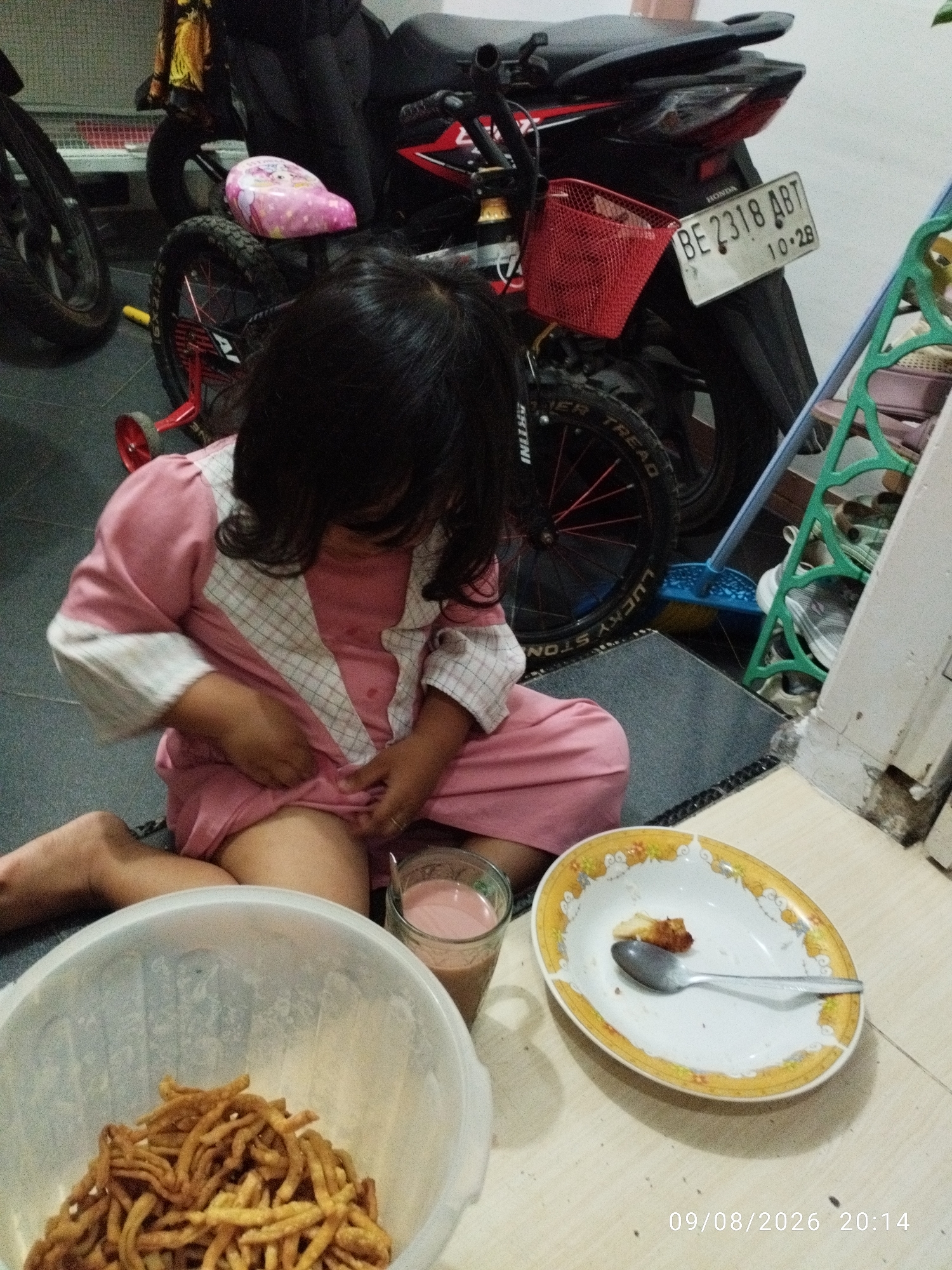 A young Indonesian child, likely a girl given her attire, is seated on a mat indoors, near a motorcycle and a bicycle. The child is wearing a pink garment with white checkered detailing on the chest and sleeves. Her dark hair partially obscures her face as she looks down.
In front of the child, there's a large white bowl filled with what appears to be fried snacks, possibly noodles or crackers. To the right of the bowl, a glass contains a pinkish beverage, likely milk or juice. Next to the glass is a shallow oval plate with a yellow and white floral pattern, containing a single piece of food and a spoon.
The background reveals a motorcycle, a bicycle with training wheels, and various household items. A license plate on the motorcycle reads "BE 2318 ABT 10-28". The flooring is tiled. A timestamp in the lower right corner indicates "09/08/2026 20:14". The scene suggests a casual moment at home, possibly in Bandar Lampung, Indonesia, given the license plate format and the presence of typical Indonesian snacks and beverages.