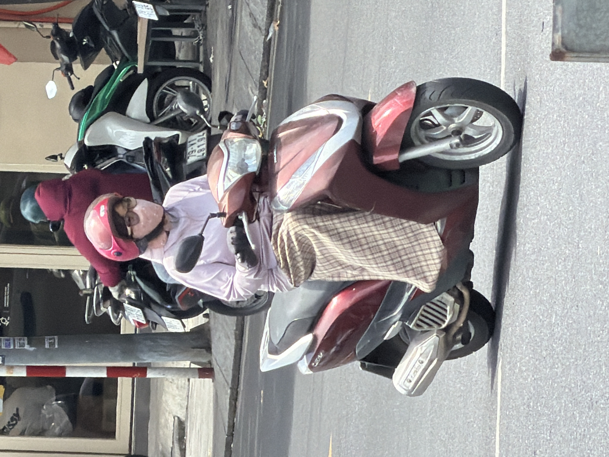A person wearing a pink helmet and glasses rides a red scooter on a street in Nha Trang, Vietnam. The person is wearing a light purple shirt and a plaid skirt. In the background, other scooters and people are visible. The street appears to be paved with asphalt, and there is a sidewalk on the right side of the image. The overall scene suggests a typical day in a bustling Vietnamese city.