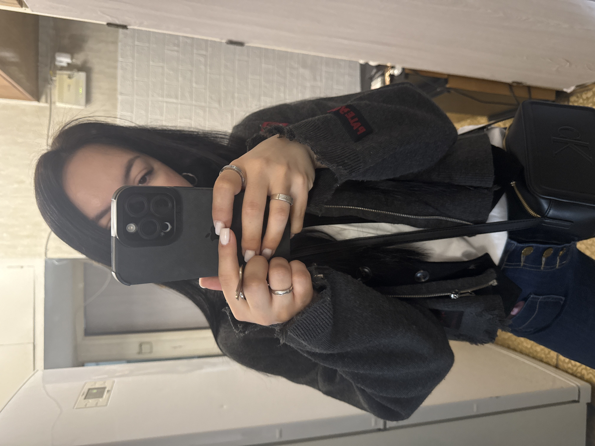 A person is taking a selfie in front of a mirror. They are holding a black smartphone in their left hand. The person has long, dark hair and is wearing a gray jacket with red text visible on the sleeve, a white top, and blue jeans. A black Calvin Klein bag is also visible in the frame. The background includes a white brick-patterned wall and what appears to be a kitchen or bathroom area, with white cabinets and tiles. The location is likely indoors, but the exact city and surrounding environment are not apparent from the image, thus it is not possible to determine it is Mytishchi, Russia.