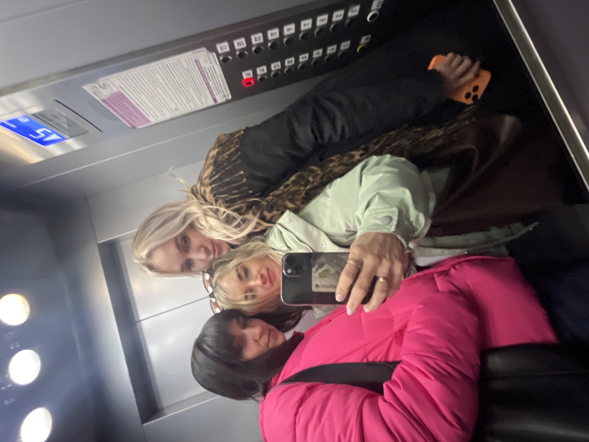 This image captures a selfie of three women inside a reflective elevator in Izhevsk, Russia. The women are seen in reflection, smiling and posing for the camera.
In the foreground, a woman with dark hair wears a bright pink puffer jacket and has a black shoulder strap visible. Next to her, a blonde woman in a light green jacket holds a black smartphone, seemingly taking the selfie. She wears glasses perched on her head and a gold-colored ring on her left hand. Another blonde woman, wearing a patterned coat (possibly leopard print) and a dark top, stands behind and to the left of the central woman, also looking towards the camera. She holds a smartphone with a bright orange case.
The background reveals the interior of the elevator. To the left, several round, bright lights are visible, indicating the elevator's illumination. Above the women, a control panel is mounted on the wall. It features a digital display showing "ST" and "5," likely indicating the current floor or destination. Below this display, a long row of numbered buttons is visible, ranging from "B" (Basement), "P1", "P2" (Parking levels), and then from "1" up to "23." One button, "20," is illuminated red, suggesting it has been pressed or is the current floor. A white document with purple accents, possibly an elevator information sheet or certificate, is affixed to the panel. The metallic and mirrored surfaces of the elevator reflect the light and the subjects. The overall atmosphere appears casual and friendly.