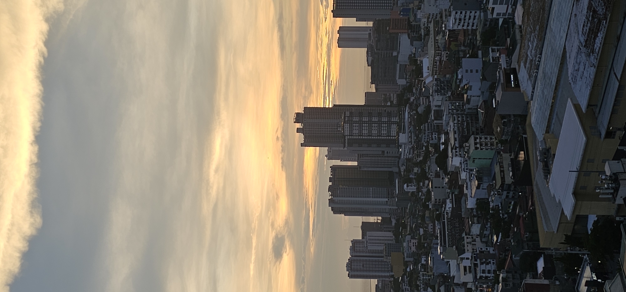 The image shows a cityscape of Makati, Philippines, at dusk. The sky is filled with a gradient of orange and yellow hues, with some wispy clouds. In the foreground, there are numerous buildings of varying heights and architectural styles, ranging from residential apartments to commercial high-rises. The buildings are densely packed, characteristic of a major urban center. Some rooftops are visible, revealing flat surfaces with some exposed utilities and architectural features. The overall scene depicts a bustling metropolitan environment under a warm, atmospheric sky. There are no visible people or specific actions taking place within the frame. The lighting suggests a transition from day to night, with artificial lights beginning to illuminate some of the structures.