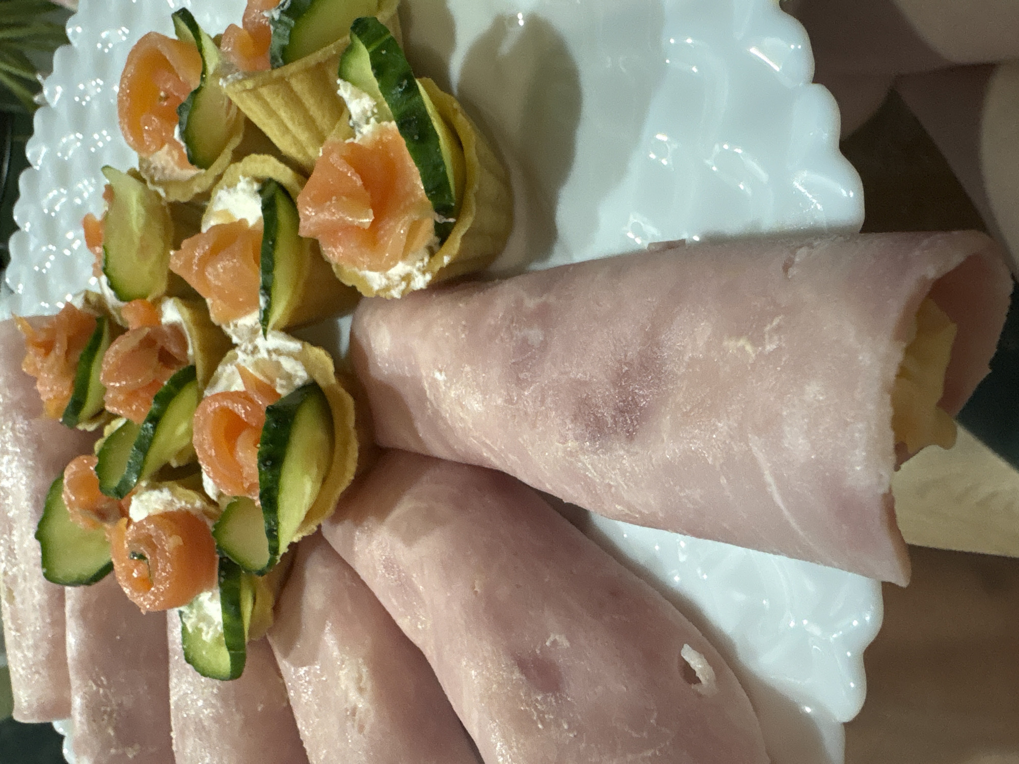 A close-up shot shows a white scalloped-edge plate displaying appetizers. On one side are several mini tartlets filled with cream cheese, salmon, and cucumber slices. On the other side, several slices of rolled-up ham are arranged. The setting does not clearly indicate any specific location within Chaikovsky, Russia.