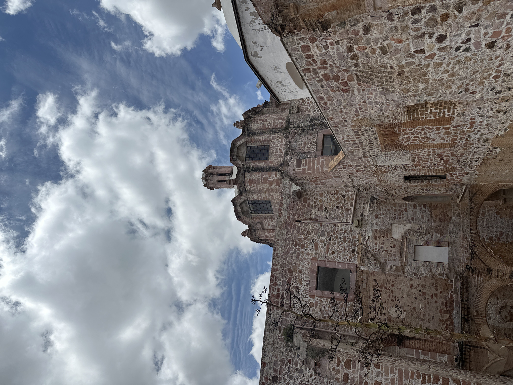 The image shows the exterior of a stone building, likely a historic church or convent, in Zacatecas City, Mexico. The building is constructed of rough-hewn stones of varying reddish-brown hues. In the foreground, to the left, a tall, leafless plant with a thick stalk and intricate branches stands against the stone wall. The plant appears to be an agave or a similar succulent.
The building has a rustic, aged appearance. Windows with dark panes are visible, some set into arched openings. Some sections of the wall show signs of decay or incomplete construction, with exposed brickwork in places.
The dominant feature of the building in the center of the frame is a domed structure topped with a lantern, reminiscent of a church cupola. The dome is partially obscured by the surrounding stone architecture. To the right, a white structure, possibly a more recent addition or a well-preserved section of the original building, contrasts with the stone.
The sky above is filled with dramatic white and gray clouds against a bright blue sky, suggesting a dynamic weather day. The perspective is looking upward, emphasizing the scale and architecture of the building. The overall impression is one of historical significance and architectural grandeur, set against a backdrop of natural beauty and an ever-changing sky.