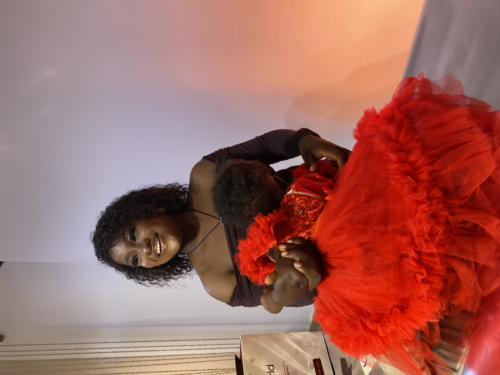 In Gura Top, Nigeria, a woman with dark curly hair and a beaming smile embraces a baby. The woman wears a dark, off-the-shoulder dress, while the baby is dressed in a vibrant red, layered tulle dress. They are positioned in an indoor setting with a plain, gradient-colored wall behind them, transitioning from white to pale orange. To the left, a white paper bag with text is visible. The lighting suggests a professional or staged photograph, possibly a studio or a well-lit event space.
