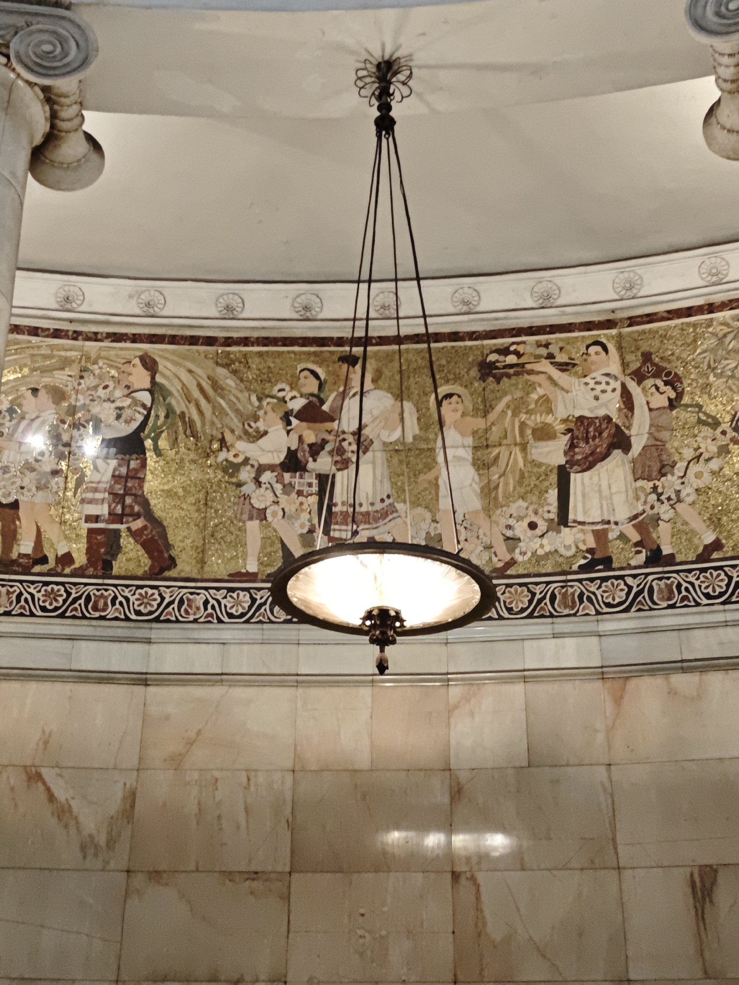 The image shows a mosaic frieze depicting people in traditional clothing carrying flowers and produce. A chandelier hangs from the ceiling, illuminating the scene. The mosaic is set in a curved wall with marble paneling below. The figures in the mosaic are rendered in a stylized manner, with a focus on their clothing and the items they are carrying. The overall impression is one of a harvest festival or a celebration. The location is unknown.