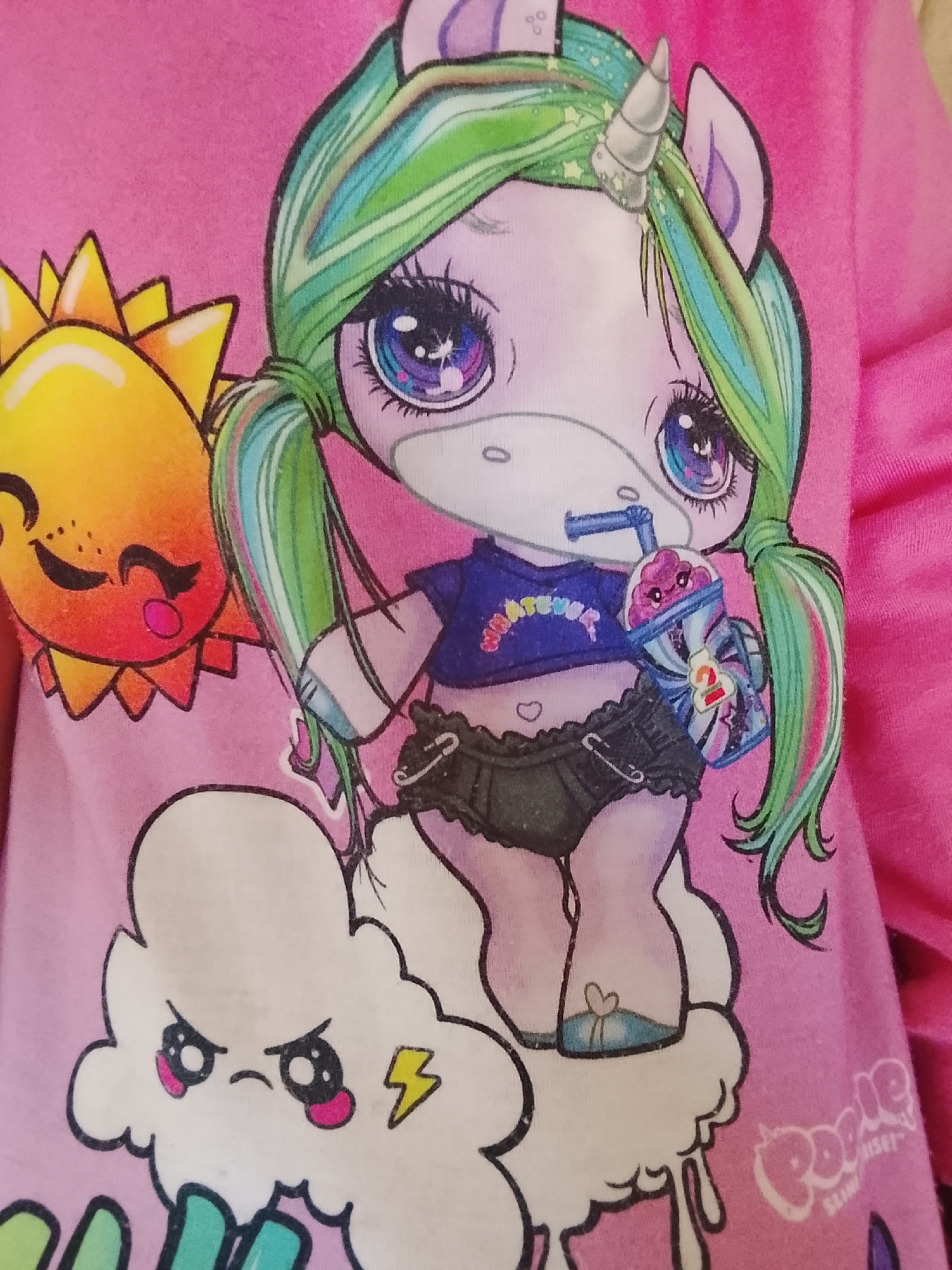 A close-up of a pink shirt with a cartoon graphic design. The central figure is a stylized unicorn with green and multicolored hair styled in pigtails, large eyes, and a silver horn. The unicorn is depicted on a white cloud with a frowning face and a lightning bolt symbol. The unicorn is holding a beverage with a straw and the number "2" on the container. The unicorn wears a dark blue cropped shirt with a rainbow-colored "WHATEVER" text and dark shorts. A cartoon sun with a face and a tongue sticking out is on the left side of the image. The words "Poopsie Slime Surprise" are on the lower right. No location of Altagracia, Venezuela, is detectable within the image.