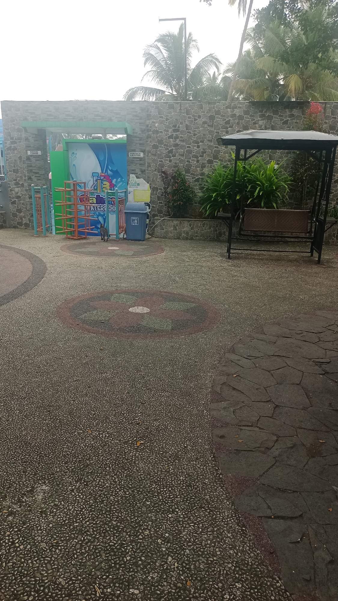 The image shows an outdoor scene in Pakemitan, Indonesia, likely a recreational area, with a stone wall in the background. In front of the wall, there is a doorway or entrance with bright graphics on the wall, featuring blue and white waves and the text "WATER BOO". To the left of the entrance, there are colorful metal structures, possibly playground equipment. To the right, a blue trash can with a recycling symbol is visible. Further to the right, a dark metal swing set with a woven seat is positioned under a canopy. Lush green plants are growing behind the swing set and around the base of the wall. The ground in the foreground is paved with small, light-colored pebbles, with two circular decorative patterns embedded in it. A larger, darker stone pathway is visible on the right side of the image. A grey cat is standing on the pebble pathway near the entrance. The sky is overcast, and palm trees are visible above the stone wall, suggesting a tropical or subtropical climate.