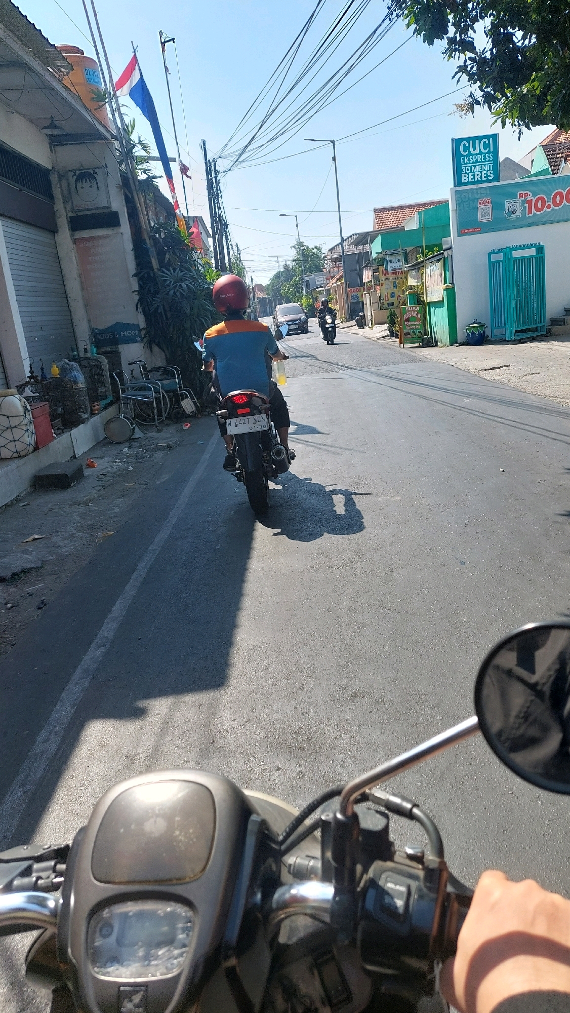 A first-person perspective shows a motorcycle's handlebars in the foreground, with a hand gripping the right handlebar. The rider is looking forward down a paved street lined with buildings and trees, characteristic of Sidoarjo, Indonesia. The street is occupied by other vehicles, including a motorcycle with a rider wearing an orange and blue shirt and a red helmet. A car is also visible further down the road. The sky is clear and blue, indicating a sunny day. A prominent sign for "CUCI EXPRESS 30 MENIT BERES" (Express Laundry, Finished in 30 Minutes) with a price of Rp. 10.000 is visible on the right side of the street. The architecture of the buildings, the presence of flags, and the style of the vehicles are consistent with the urban environment of Sidoarjo.