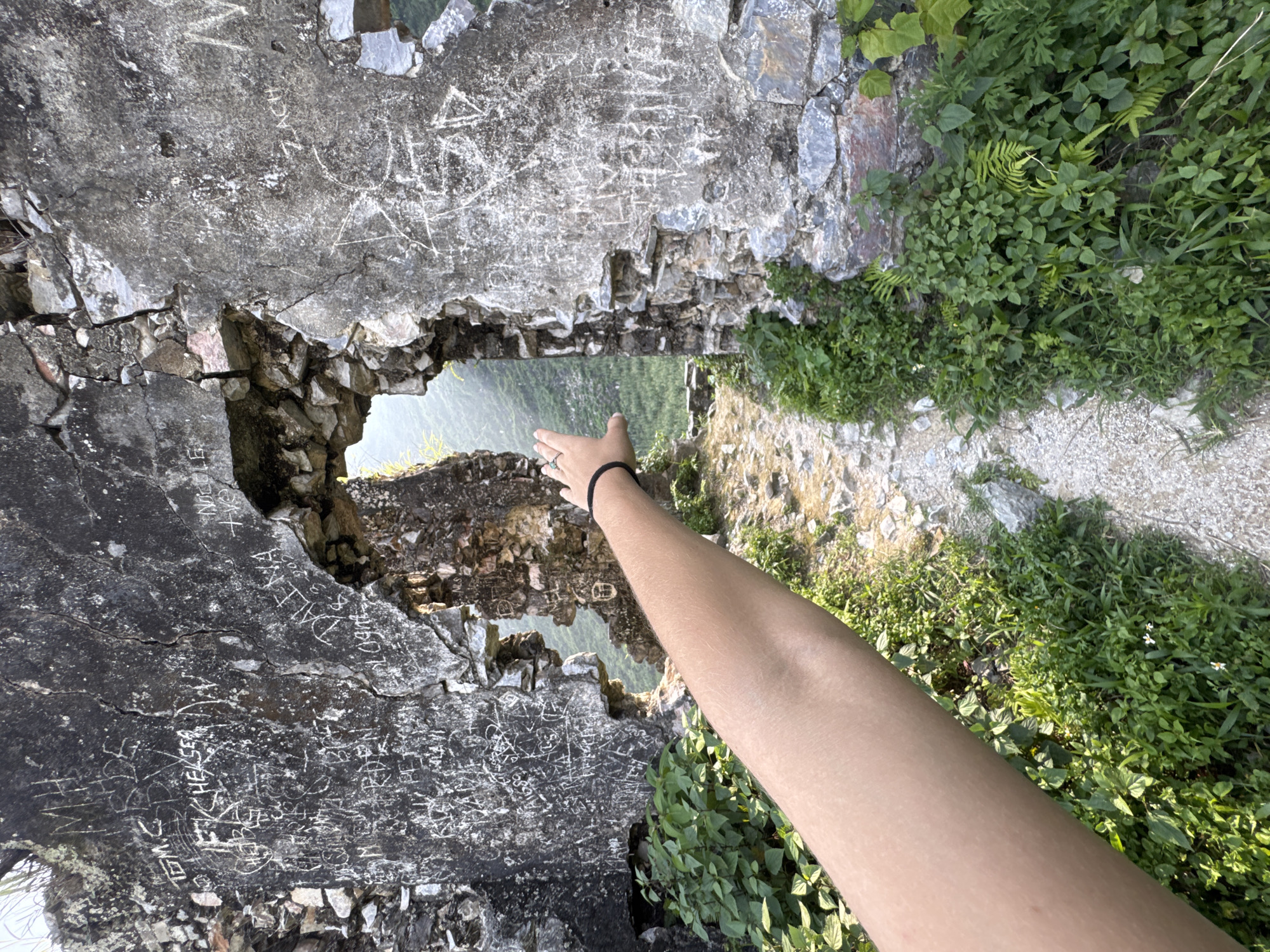 The image depicts a section of ruined stone wall in Đường Thượng Commune, Vietnam. Graffiti covers the wall's surface, including names like "ZACH" and illegible phrases. A hole in the wall provides a glimpse of a distant landscape. An arm reaches into the frame from the lower right, the hand extended toward the opening. The ground on the other side of the wall is covered in vegetation and a path.