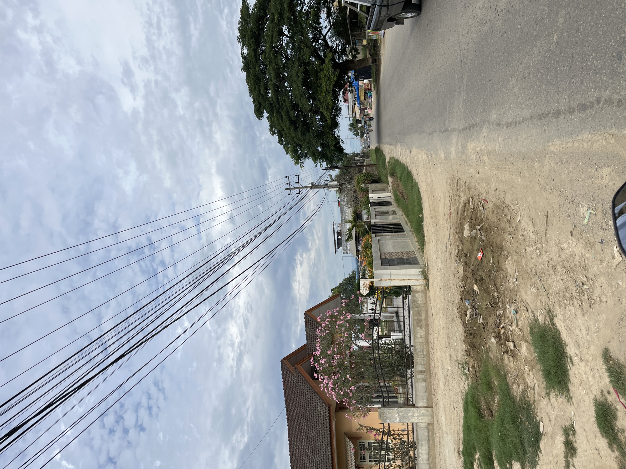 The image captures a street scene in Landuh, Indonesia, on a cloudy day. In the foreground, a dirt and gravel area with patches of grass and scattered debris is visible on the left side. A residential property is bordered by a fence, featuring a house with an orange-toned exterior and a brown tiled roof. A vibrant pink flowering tree is prominent in the yard. The street itself is paved and curves to the right. On the right side of the street, a large, mature green tree with a wide canopy dominates the landscape. Further down the road, there are small buildings, some with blue and red awnings, and several motorcycles are visible, indicating local activity. Power lines are strung across the sky, supported by utility poles. A black pickup truck is parked on the right side of the road near the foreground.