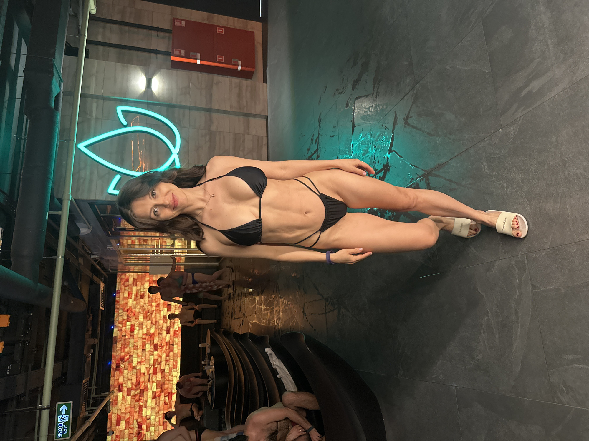 The image captures a woman in a black bikini posing in a spa setting. She is lying on a dark tile floor, wearing sandals. In the background, there is a group of people sitting or lying down inside an area with a wall covered in red brick-like tiles, suggesting a salt room or similar relaxation space. Above the woman is a wall with a bright blue neon art installation, a fire extinguisher is mounted on the wall, and what appears to be a pipe. The lighting suggests a controlled, indoor environment with specific accent lighting, like the turquoise glow. Given the context, it is implied that this is a spa or sauna facility, possibly in Saint Petersburg, Russia.
