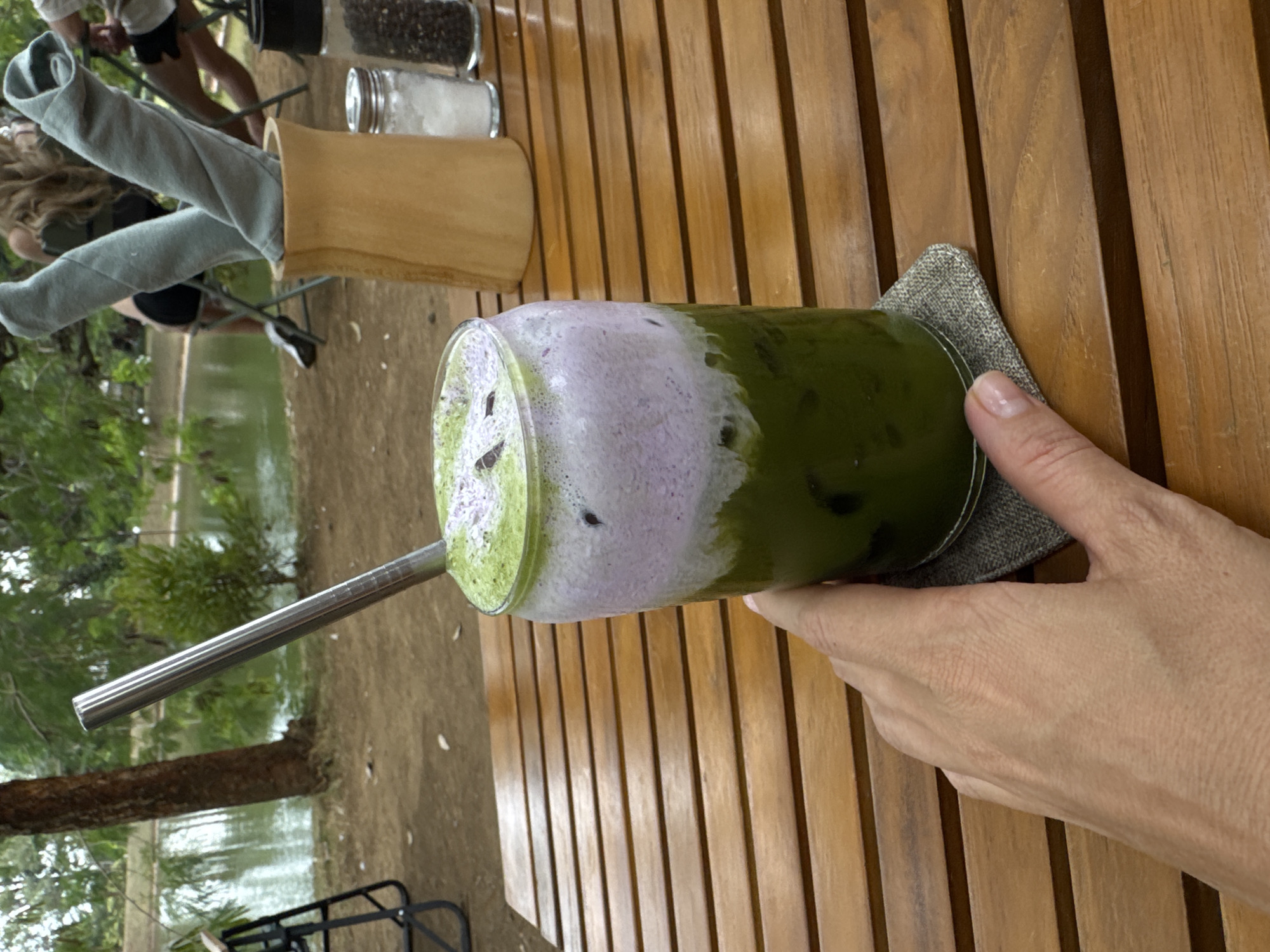 A hand holds a tall glass of layered green and purple iced beverage with a metal straw, resting on a wooden table. In the background, a body of water is visible, surrounded by trees and people seated at tables. The scene appears to be an outdoor cafe or restaurant in Kuta Selatan, Indonesia. The beverage has a frothy purple layer on top of a green liquid with ice. A grey coaster is under the glass. To the right, a wooden holder contains rolled grey napkins, and behind it are salt and pepper shakers. The table is made of horizontal wooden planks. The overall atmosphere suggests a relaxed, tropical setting.