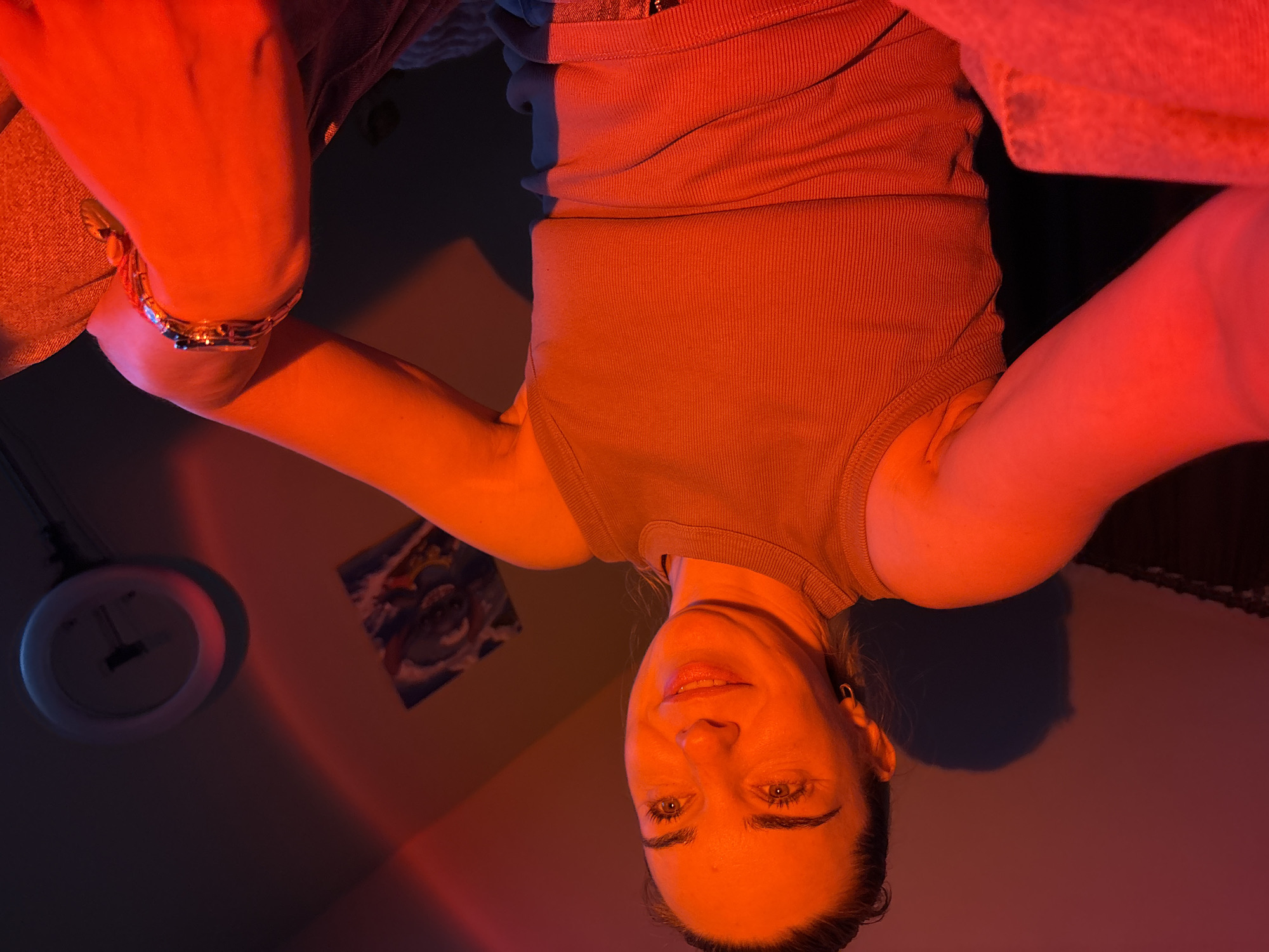 A medium shot shows a woman in a sleeveless, ribbed, tan top illuminated by red lighting, casting a warm glow on her skin and the surrounding room. Her head is turned to the right, and she is looking upwards with a slight smile. She wears a gold hoop earring in her left ear and a gold wristwatch on her left arm. Her right arm is bent, resting on her chest. In the background, a circular clock with a black face and white numbers is mounted on the wall. To the left of the clock, a framed picture depicting a creature is visible. The room appears to be a bedroom, with soft furnishings and a wall with a subtle shadow pattern cast by the lighting. The overall ambiance is intimate and artistic, amplified by the dramatic red lighting. The location is Unknown, Russia.