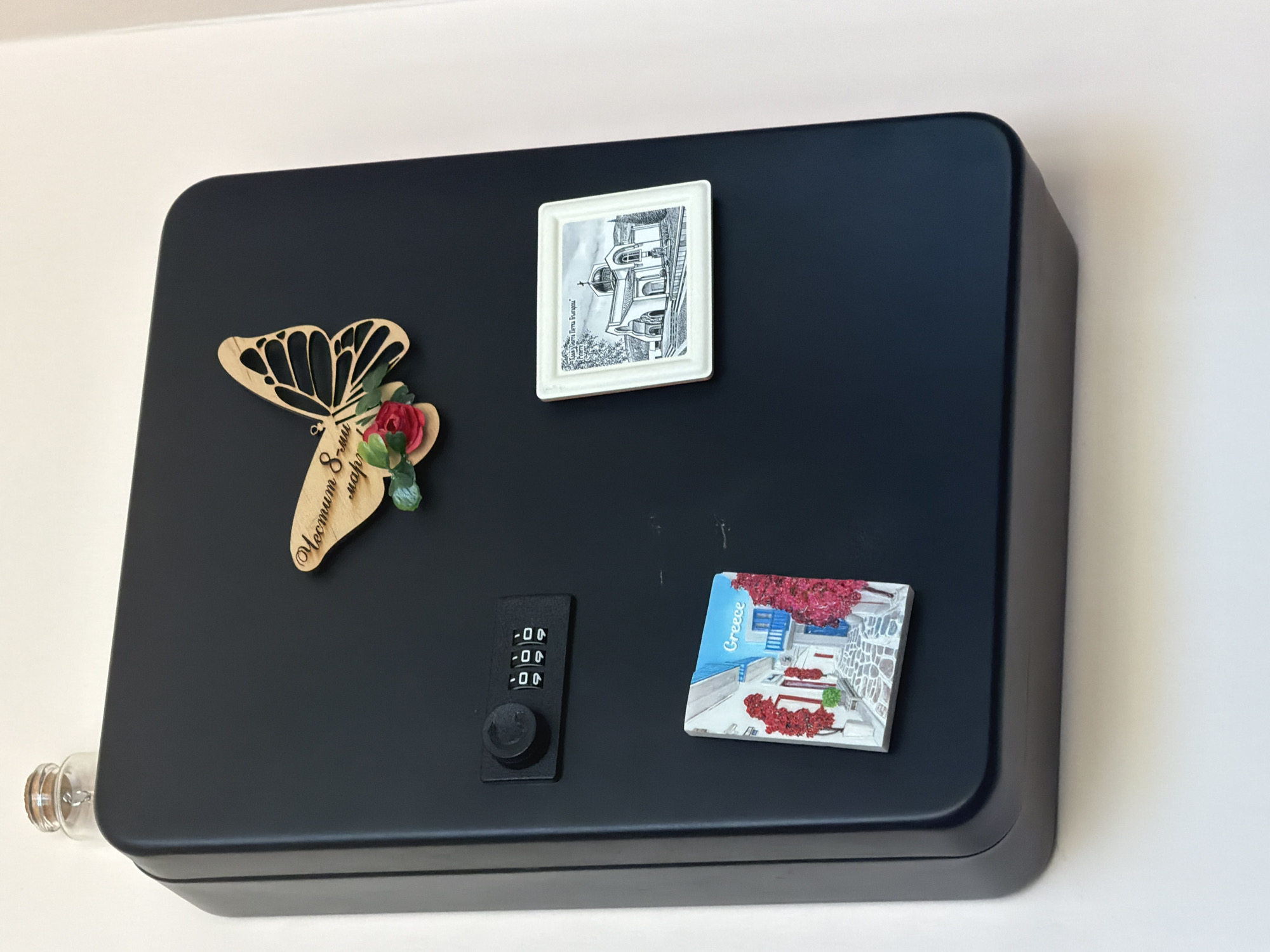 The image shows a black, rectangular lockbox mounted on a white wall. On the top of the lockbox is a small glass bottle with a cork stopper. Attached to the lockbox are three decorative items: a wooden butterfly with flowers and text, a small square magnet depicting a street scene in Greece with the word "Greece" printed on it, and a square magnet featuring a black and white drawing of a church, likely in Bulgaria, with text above it. The lockbox also has a combination lock. The scene is brightly lit, and the background is a plain white wall. There is no indication of the city of Dolno Ablanovo, Bulgaria.