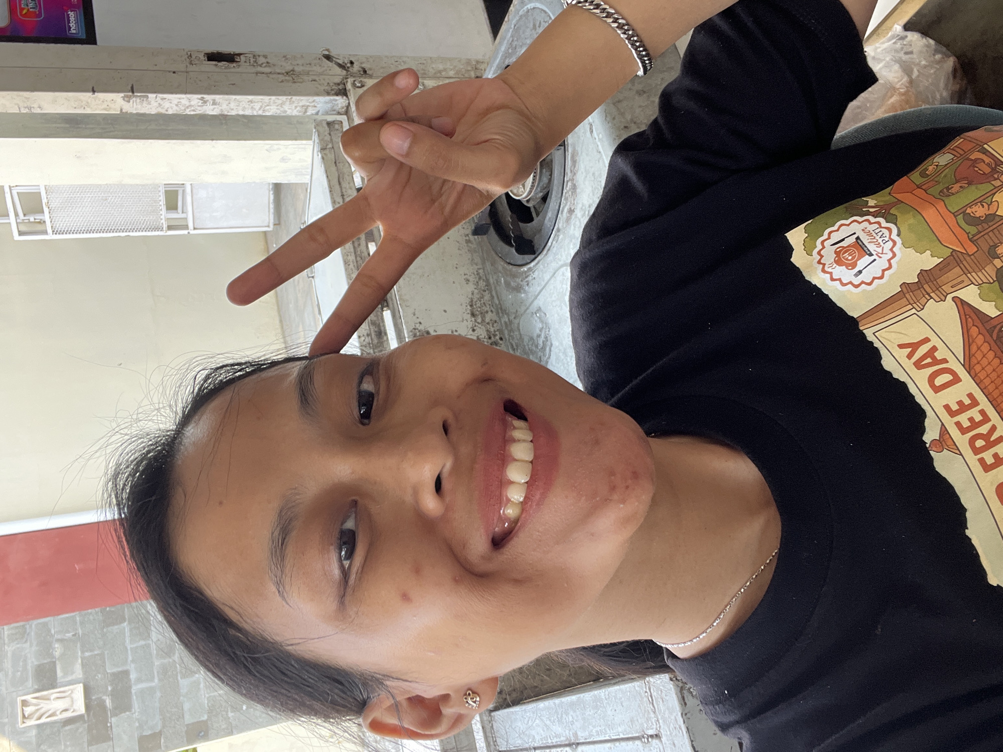 A young woman with dark hair and brown eyes smiles broadly at the camera, holding up two fingers in a peace sign. She wears a black t-shirt with a graphic design on the front. The background appears to be an outdoor or semi-outdoor area, possibly a street or an open-air market. There are brick walls, a white fence or gate, and some furniture visible. The lighting suggests it is daytime. The image is a close-up, focusing on the woman's face and upper body.