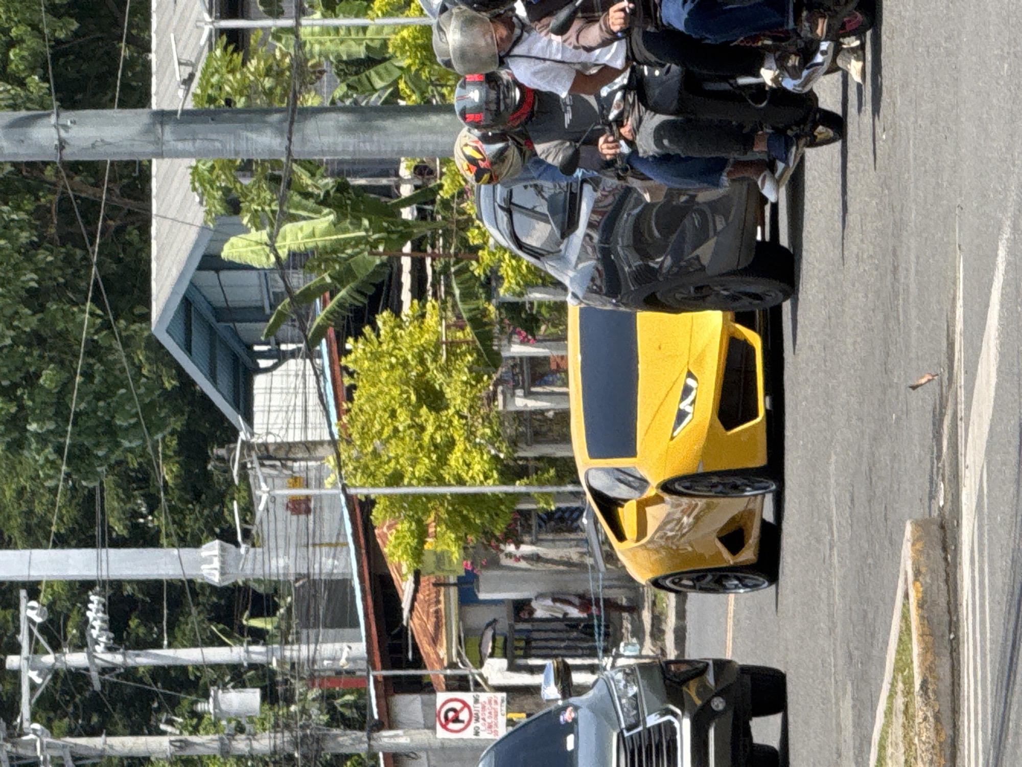 The image shows a bright yellow sports car, likely a Lamborghini, stopped at a street intersection in Davao City, Philippines. The car is positioned in the foreground, angled slightly towards the viewer. To its left is a dark colored SUV. To the right of the sports car, a group of people are riding on a motorcycle, also stopped at the intersection. They are wearing helmets, with one person in the front and two passengers behind.
The environment suggests a busy urban street. In the background, there are residential buildings with corrugated metal roofs, lush green trees, and utility poles with numerous wires. One building has a sign with a red circle and a diagonal line, indicating a restriction. A person in a white shirt and red shorts can be seen standing near a gate on the left side of the image, between the SUV and the yellow car. The road surface is asphalt, with painted lines indicating traffic lanes and a concrete divider. The lighting indicates it is daytime, with bright sunlight casting shadows.