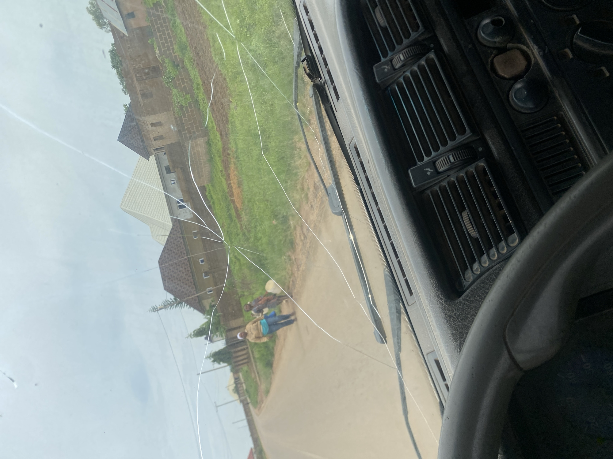 The image shows a view from inside a vehicle, looking through a cracked windshield at an outdoor scene. The dashboard of the vehicle is visible in the foreground, with parts of the steering wheel and air vents. Outside, a paved road curves to the left, bordered by a grassy verge. Two individuals, appearing to be women, are walking along the road. One is wearing a tan jacket and blue pants, and the other is wearing a patterned dress. Behind them, there are several buildings with brick walls and varied roofing styles. The sky is overcast. The location is identified as DU, Nigeria.