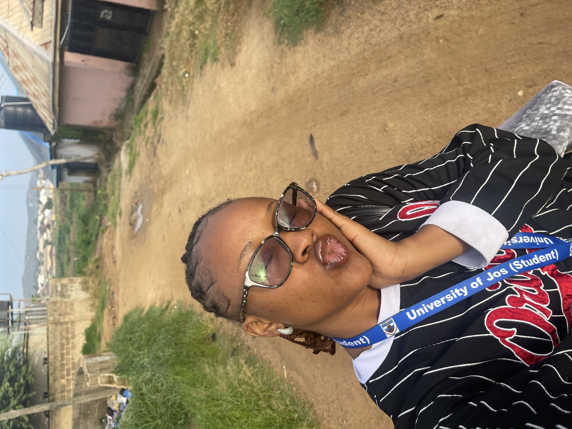 A young woman is taking a selfie outdoors in a rural setting in Rafin Par, Nigeria. She is wearing a black and white striped jersey and sunglasses, and has her hand on her face. She is making a kissing face. A blue lanyard with "University of Jos (Student)" written on it is visible around her neck. The background shows a dirt road, tall grass, and buildings. The time of day appears to be daytime, and the weather seems to be overcast or hazy. The overall mood of the image is casual and playful.