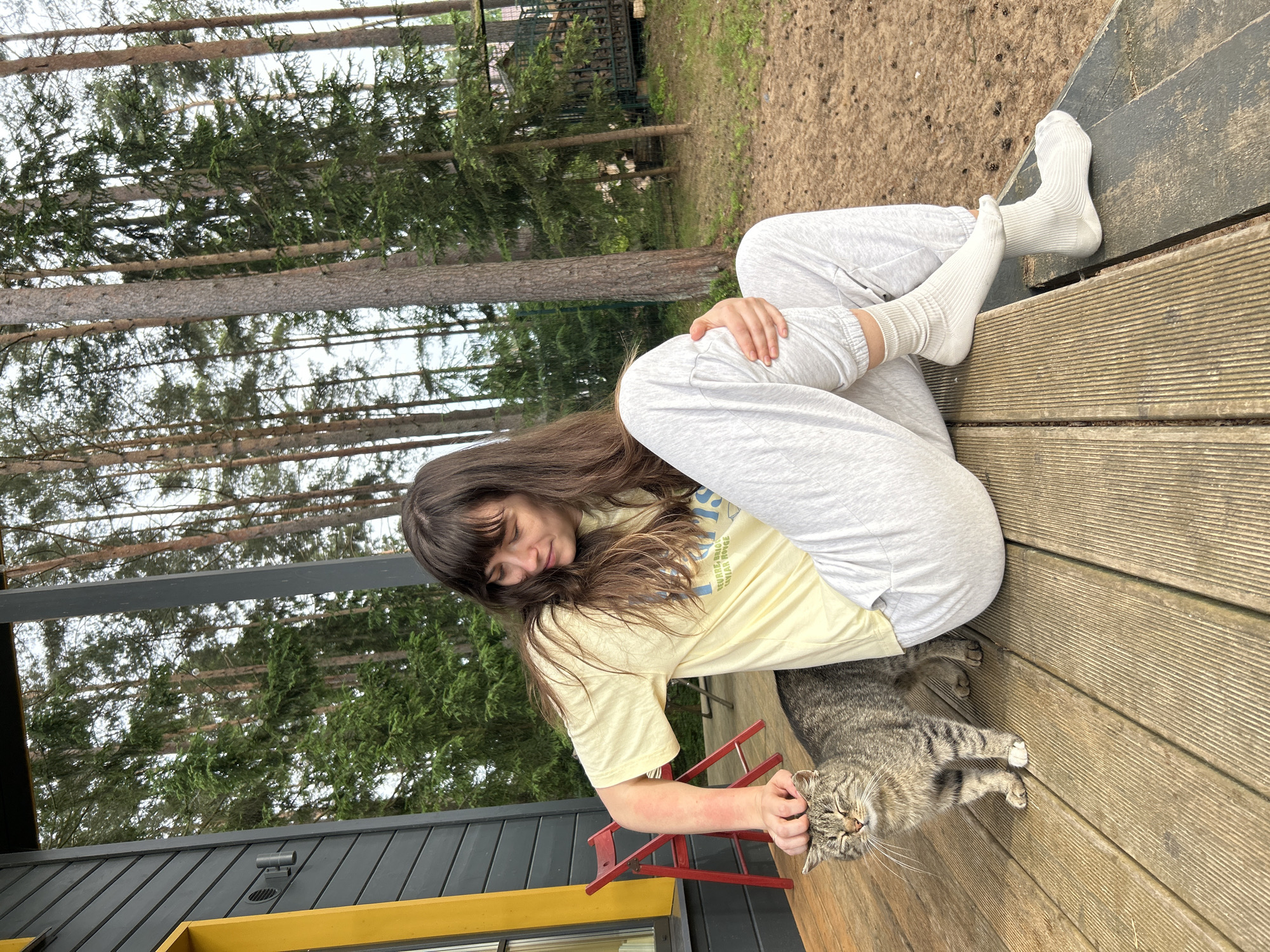A young woman with long brown hair, wearing a light yellow t-shirt and light grey sweatpants with white ribbed socks, is sitting on a wooden porch. She is petting a tabby cat that is standing next to her. The woman is looking down at the cat, who has its eyes closed and appears to be enjoying the attention. The porch is made of wooden planks and is surrounded by tall pine trees. The house to the left is dark grey with a bright yellow window frame. In the background, there is a fence and a pile of firewood. The location is the city of Shepelyovo, Russia. The overall scene is peaceful and relaxed.