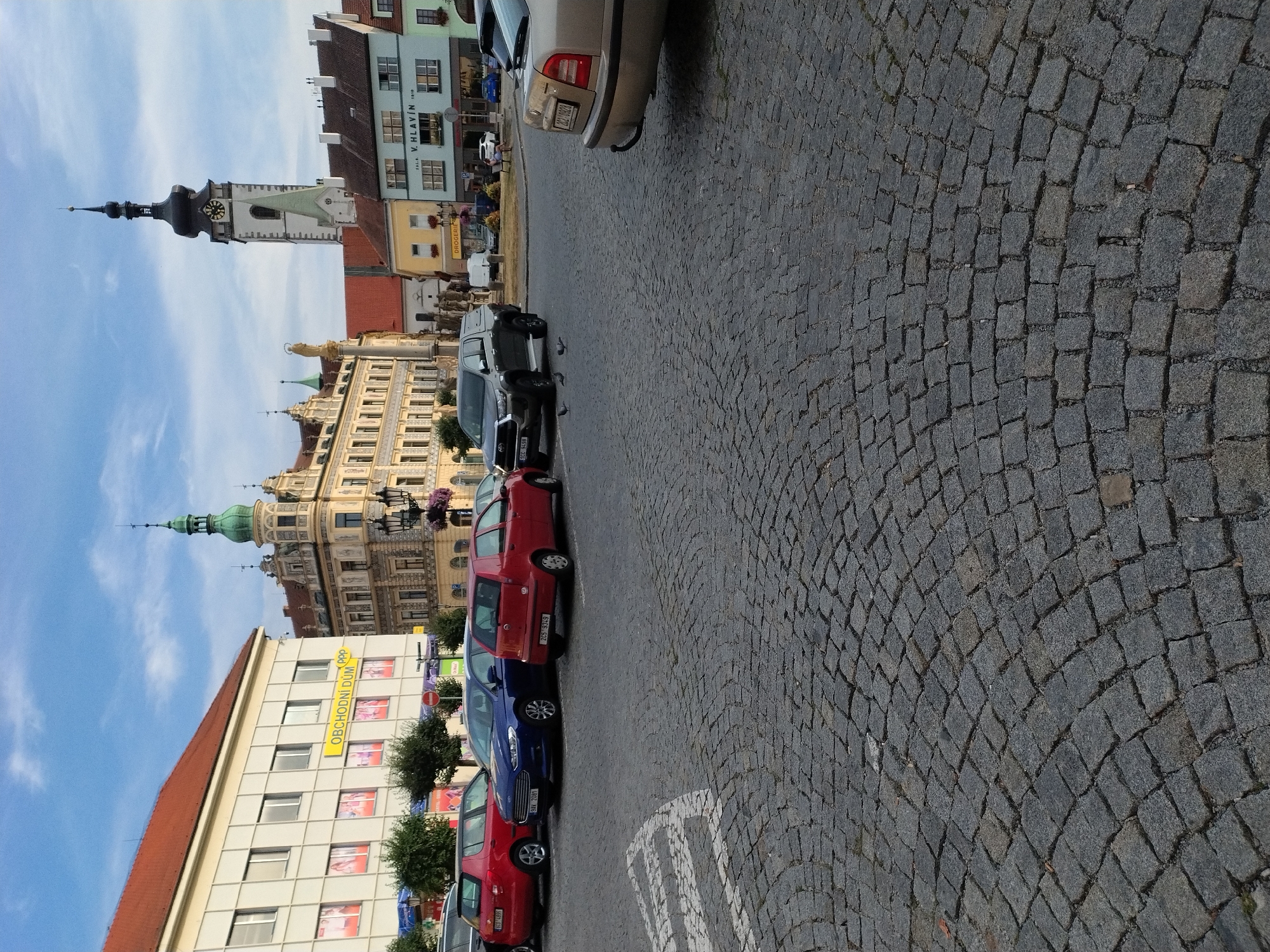 This is an eye-level, outdoor, full shot of a paved plaza in Písek, Czechia, featuring a row of parked cars and several buildings in the background. The plaza's ground is covered in cobblestones. On the left, a light-colored building with a red tiled roof has the sign "OBCHODNÍ DŮM" visible. To its right, a tan building with ornate architecture and a green-domed tower stands. Further in the background, a white church tower with a black spire and clock face dominates the skyline. Other buildings with colorful facades line the plaza. Several cars are parked: a red hatchback, a blue sedan, another red hatchback, a dark gray SUV, and the rear of a light brown car. Three pigeons are visible on the cobblestones in front of the SUV. The sky is blue with scattered clouds.