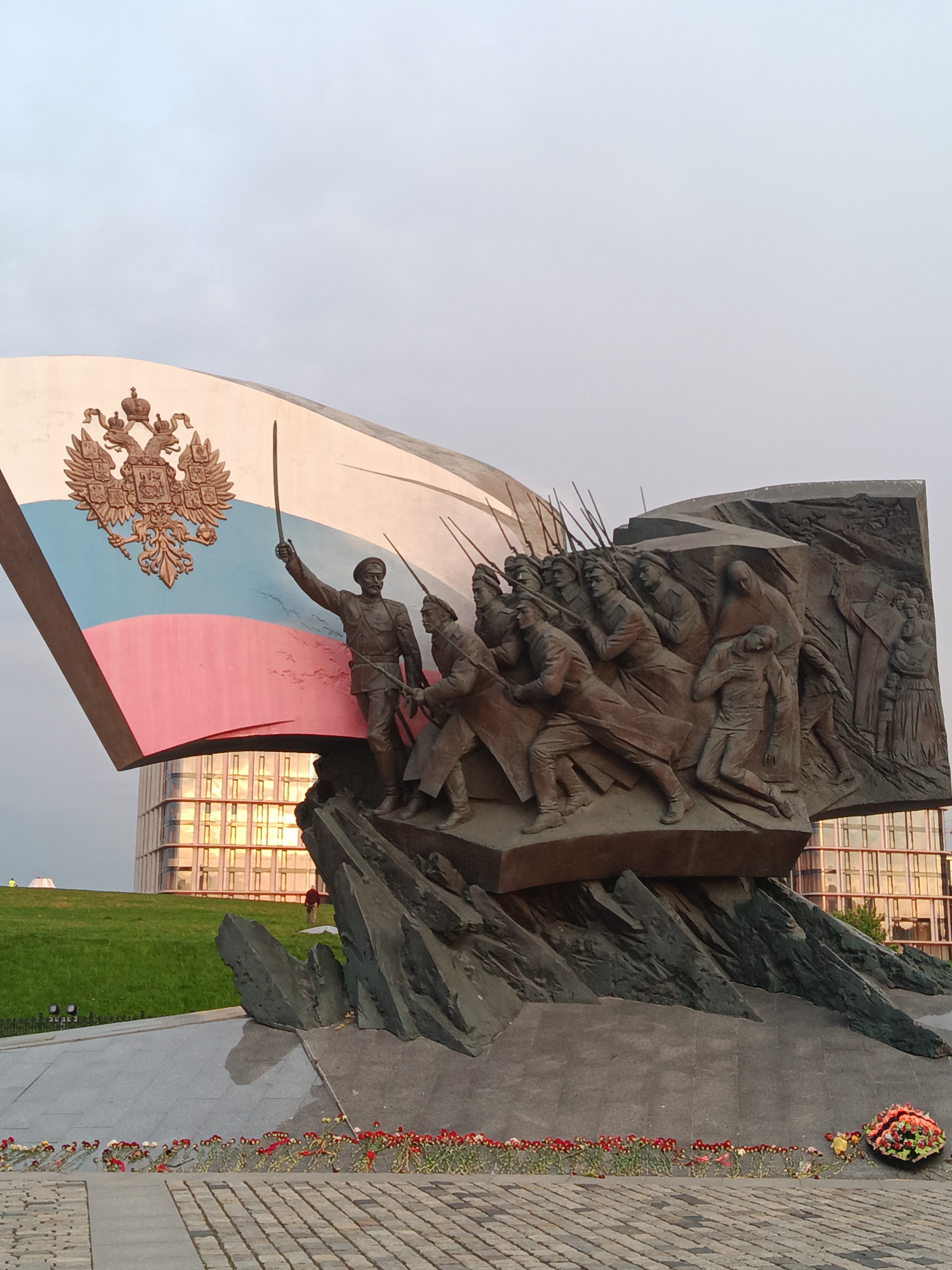 The image shows the Memorial to the Heroes of World War I, located in Moscow, Russia, on Poklonnaya Hill. The memorial features a bronze sculptural composition of soldiers in battle, emerging from a dark stone base. A large stylized depiction of the Russian flag, with the double-headed eagle emblem, serves as a backdrop to the sculpture. Flowers are laid at the base of the monument. A modern building with reflective windows is visible in the background, along with a grassy area with people, contributing to the urban setting. The sky is overcast.
