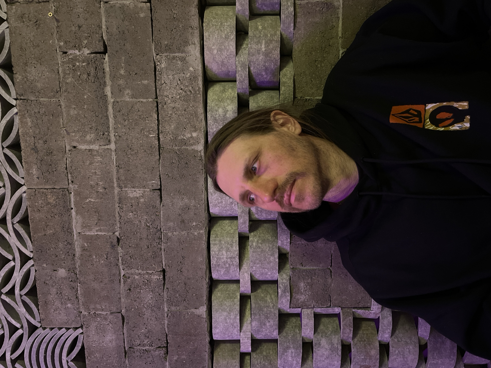 A man wearing a black hoodie stands in front of a decorative brick wall. The wall consists of gray rectangular bricks on the left and gray cylindrical bricks arranged in an intricate pattern on the right. The lighting is predominantly purple, with some ambient light. The location is likely in the city of Krasnoyarsk, Russia.