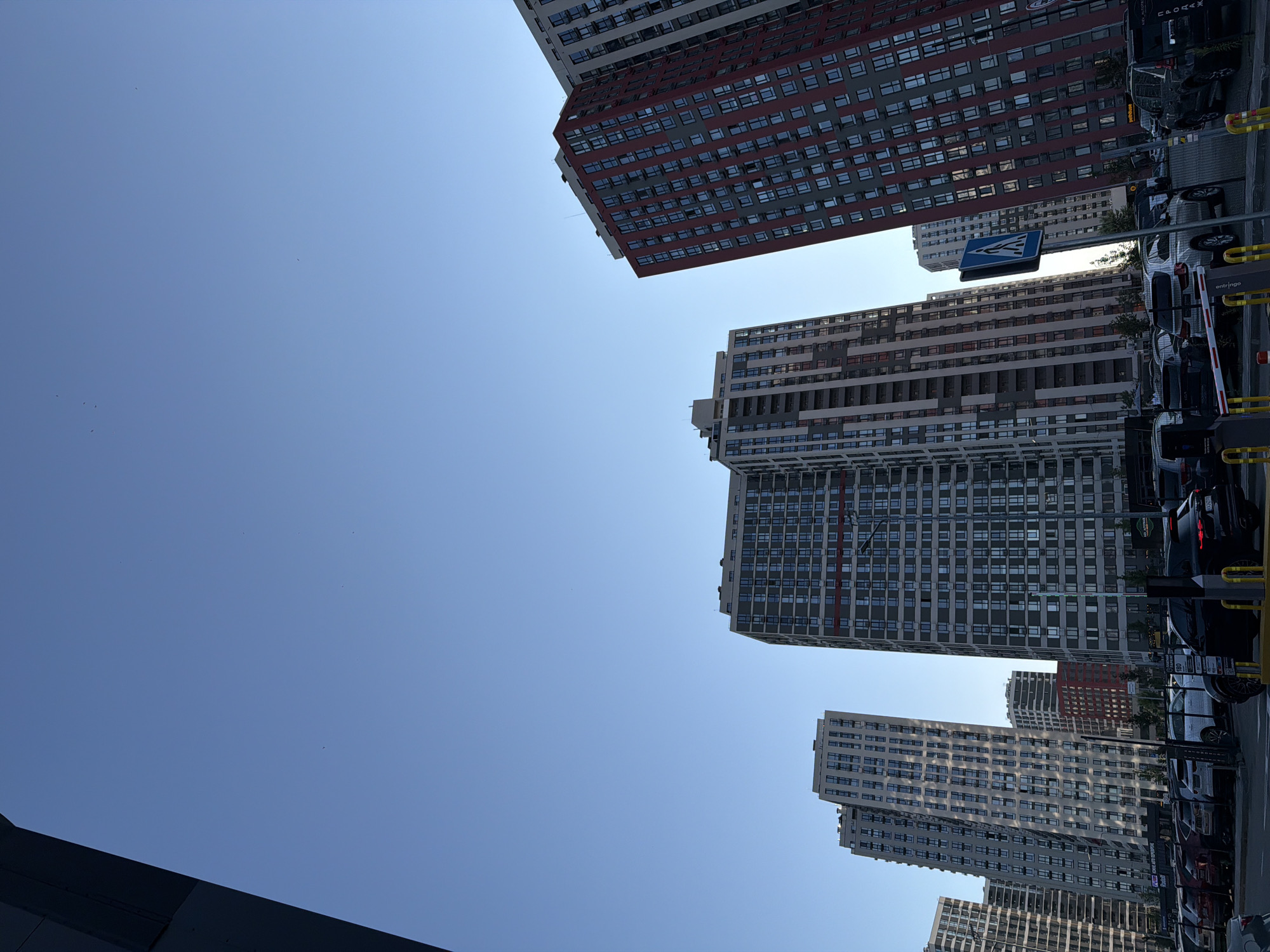 The image depicts a street-level view of several modern high-rise residential buildings in Kyiv, Ukraine, under a clear blue sky. The buildings are of varying heights and architectural styles, with some featuring prominent red and gray facades, while others are predominantly gray or light-colored. A parking lot is visible in the foreground, filled with numerous cars. Pedestrian crossings and street signs are also present. The perspective suggests the photo was taken from ground level, looking upwards at the towering structures.