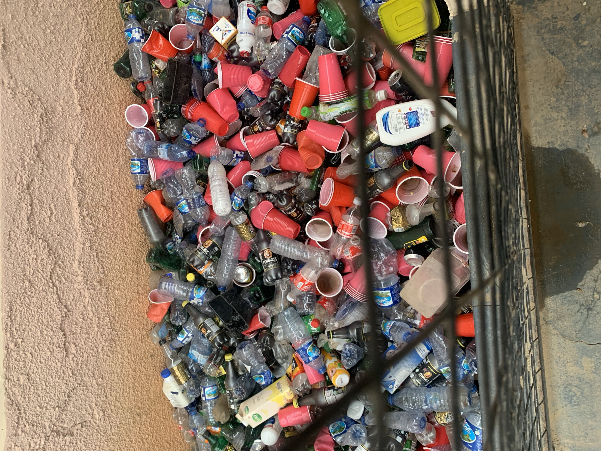 The image shows a large collection of discarded plastic bottles and red cups piled up next to a concrete surface and a rusted metal grate. Many of the bottles are branded "Swan" and "FEARLESS". The scene appears to be taken in a public or semi-public area, possibly near a stadium, given the concentration of disposable items suggestive of a large event. The light is bright, suggesting a daytime shot. Based on the identified brands, the location is likely New Stadium, Nigeria.