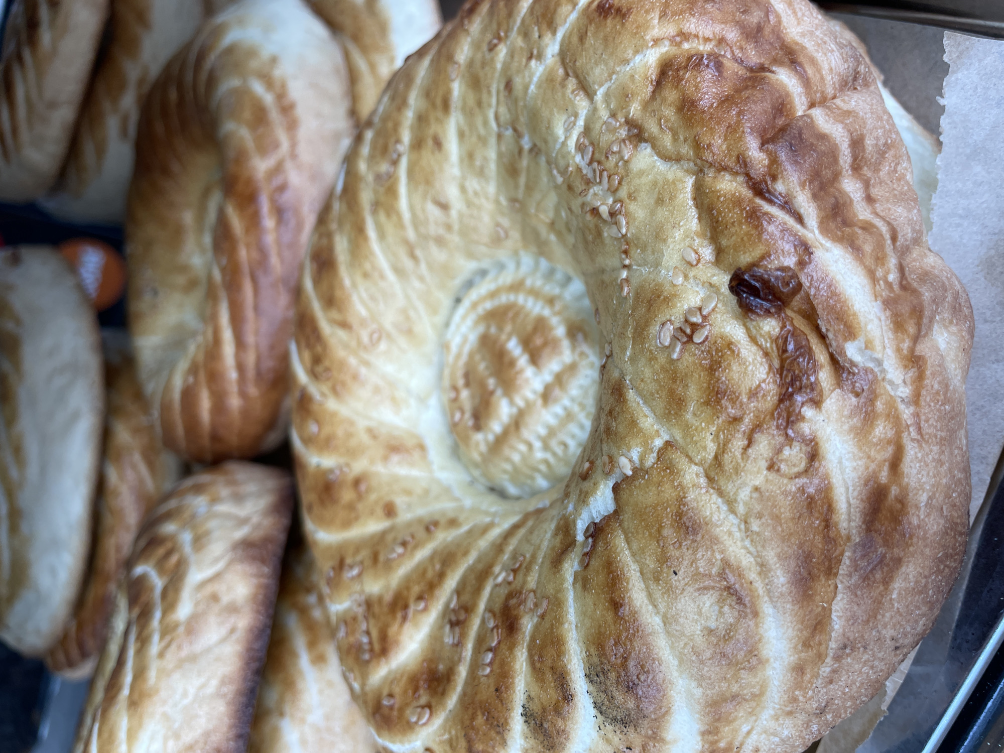 This is a close-up, overhead shot of several baked goods, likely bread, arranged together. The primary subject is a large, round, flatbread with a distinct spiral pattern pressed into its surface, emanating from a central dimple. The bread is golden brown with darker, toasted areas and is sprinkled with sesame seeds. To the left of this main bread, other similar baked goods are visible, some appearing to be elongated rolls with a similar textured crust. The baked goods are in focus, and the background is slightly blurred, suggesting a shallow depth of field. The lighting appears to be neutral, possibly from overhead. There is no visible text. The overall impression is of a food display, likely in a bakery or market.