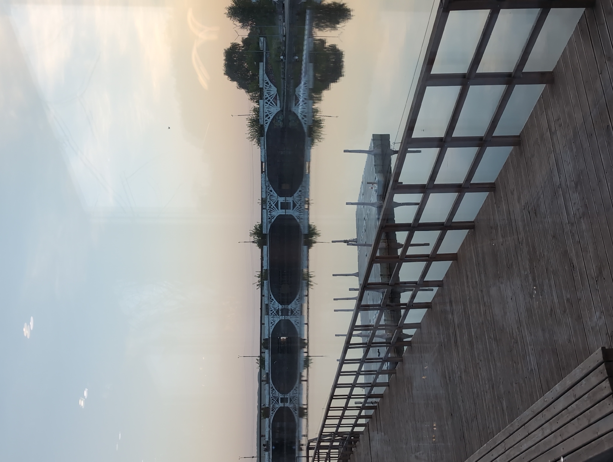 The image shows a scene in Votkinsk, Russia. A bridge is reflected in the water. A wooden structure is visible on the right side of the frame. The sky is light-colored, possibly indicating dawn or dusk. There is no visible person.