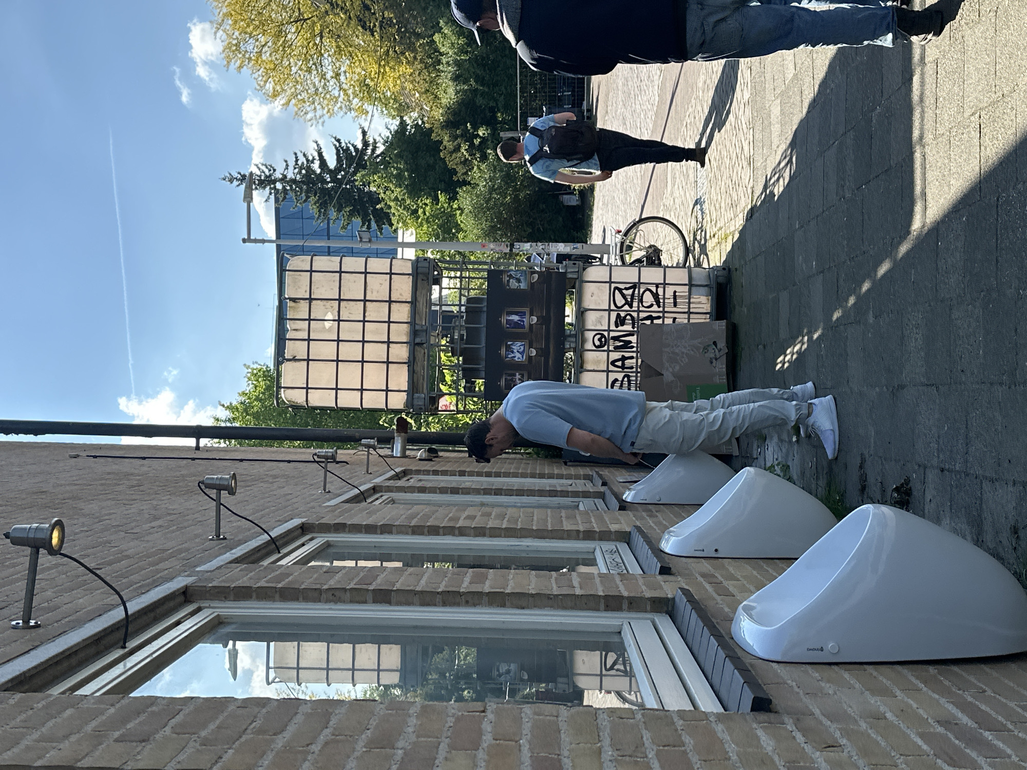 The image shows a man urinating into one of three public urinals located against a brick building, likely in Amsterdam, Netherlands, given the style of the urinals and the urban environment. The man is wearing a light blue t-shirt and khaki pants, and white sneakers. The urinals are white and egg-shaped. Above the urinals are windows with brick frames, and above the windows are outdoor lights. In the background, there are other people walking on the sidewalk, a bicycle leaning against a water tank, and trees. The sky is blue with some clouds.