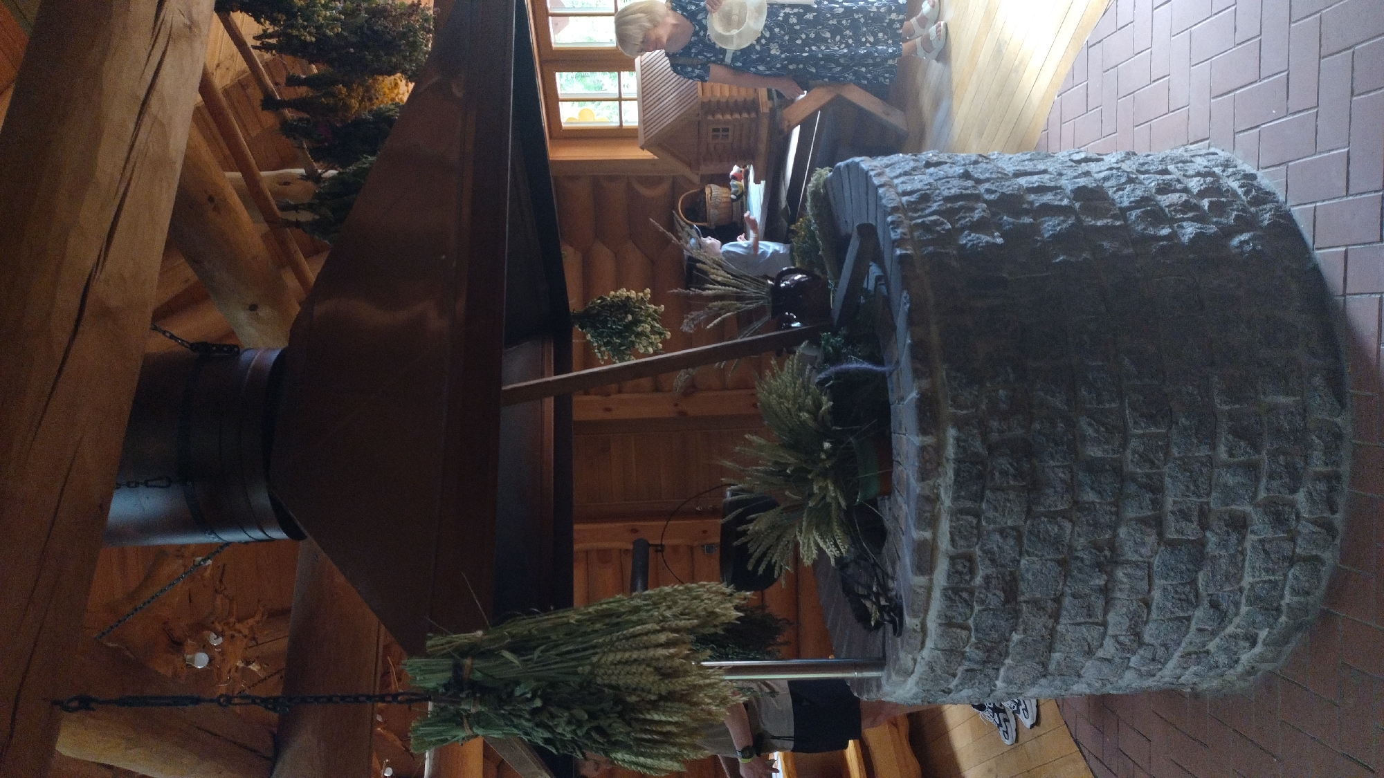 In an indoor rustic setting, a stone well with a large metal hood overhead is the central focus. Bunches of dried wheat and greenery hang from chains above and from the structure. A person on the left is partially visible, wearing a t-shirt and shorts, with sneakers. Inside the well, there are decorative plants in pots and a dark cauldron hanging from a hook. To the right, a woman in a floral dress and white sandals holds a straw hat, standing near a wooden display with a miniature house and various items. A child is also visible near the display. The environment features wooden walls and ceiling beams, and the floor is a combination of wood and brick tile.