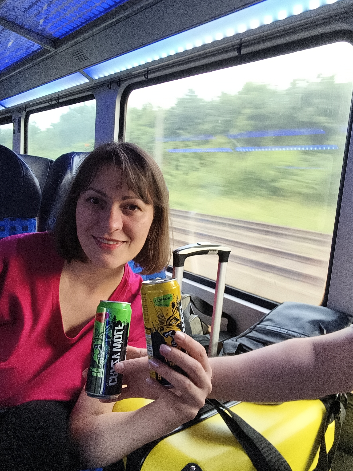 A young woman with short brown hair and bangs is smiling at the camera. She is wearing a bright pink shirt and is holding two cans of energy drinks, one green and one yellow. The green can has white text that reads "CHEEKY MOTE," and the yellow can has black and yellow text. She is seated on a train, evidenced by the blurred scenery outside the window, the train tracks, and the interior lighting of the carriage. A suitcase with a black strap is visible next to her. The overall scene appears to be a candid moment captured during travel.