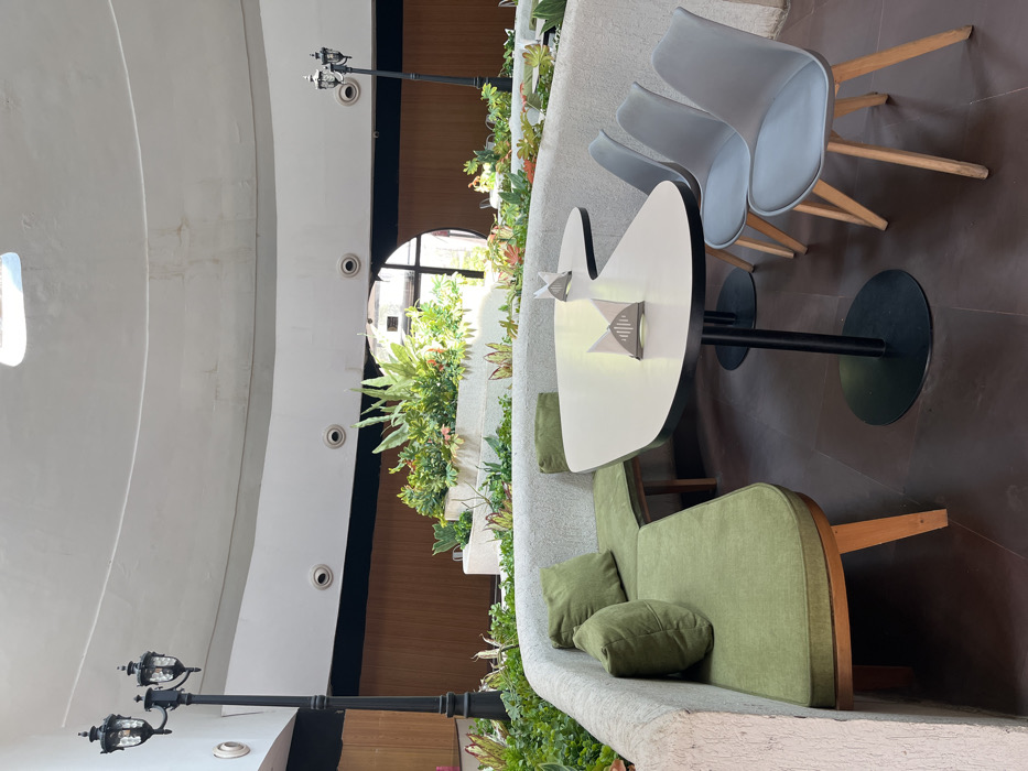 The image shows a seating area in a restaurant or cafe located in Sahibzada Ajit Singh Nagar, India, at the coordinates 30.703, 76.668. The seating area is located in a semi-circular building with a white ceiling and walls. There is a large curved green sofa with a wooden frame and two pillows. The sofa is facing a C-shaped table with four chairs. The table is made of wood and the chairs are made of metal and wood. There is a white napkin on the table. There are many plants growing in pots, lining the walls and creating a green space in the center of the room. The floor is made of a brown tile. The scene is set during the day and there are no people in the image.