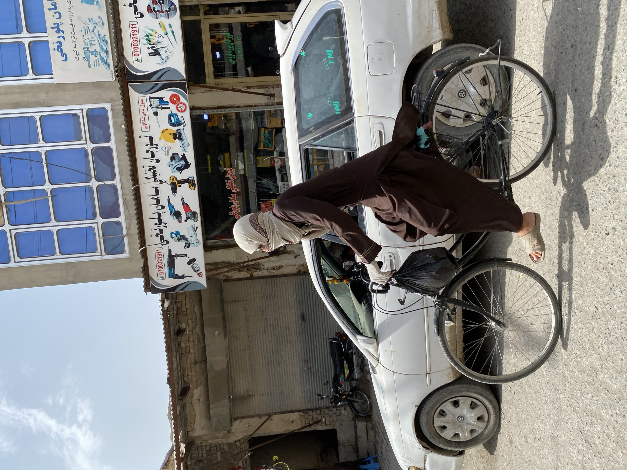 In Kandahar, Afghanistan, a person wearing a brown abaya and a light-colored headscarf rides a bicycle on a paved street. The person is wearing white gloves and carrying a black plastic bag. The bicycle has a basket filled with the bag. A white station wagon is parked next to the bicycle. The background shows a building with blue windows and several signs with text in Dari. The street is lined with shops and appears to be a commercial area. A motorcycle is parked in front of a shuttered storefront on the left side of the image. The sky is bright and clear.