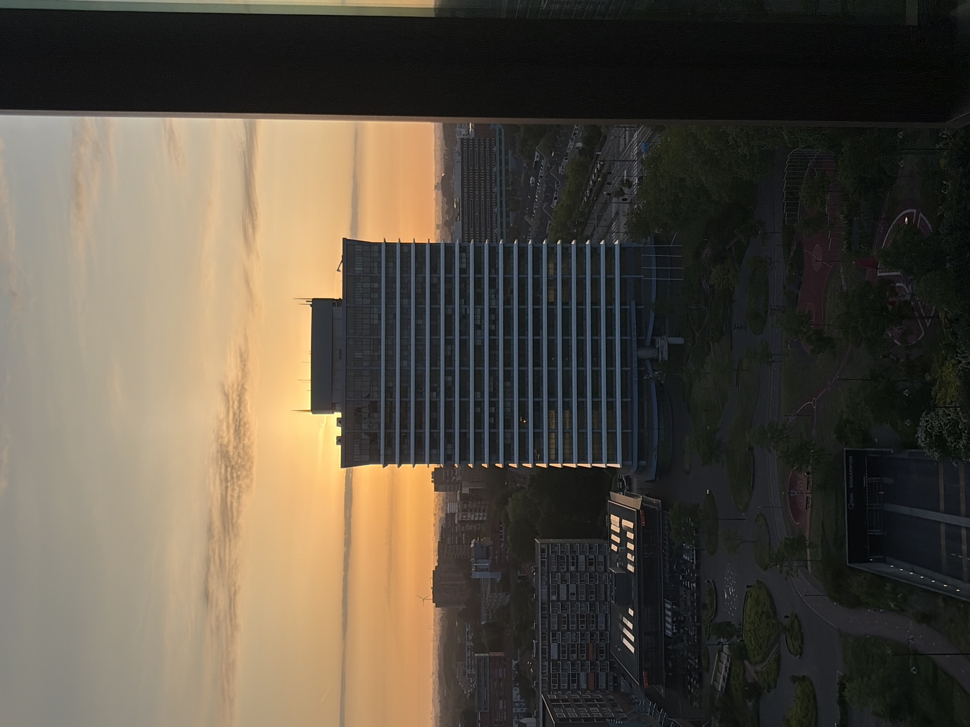 A vertical, eye-level shot looking out of a window from a high vantage point shows the skyline of Rijswijk, Netherlands, during a sunset or sunrise. The prominent building in the center of the frame is the former Shell office complex, now known as the "Leeuwenstein." To the right of the Leeuwenstein is a park with a playground and walking paths. In the foreground, the window frame is visible, indicating the photo was taken from inside a building. The sky is filled with orange and yellow hues, suggesting the time of day. Other buildings and infrastructure of Rijswijk are visible in the background, fading into the distance due to atmospheric perspective. The scene is calm and serene, with no people visibly present.
