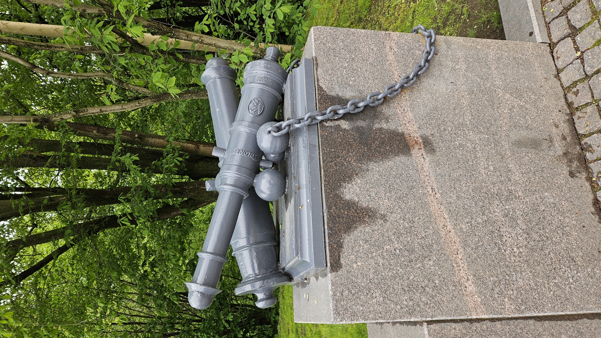 The image shows a monument featuring three gray cannons resting on a gray pedestal with a chain draped over it. The monument is located in an outdoor setting with trees and green foliage in the background. To the right of the monument is a paved walkway with small stones. The inscription "OLONETS" is visible on one of the cannons. The location appears to be in a city, but the specific city and region are unknown.