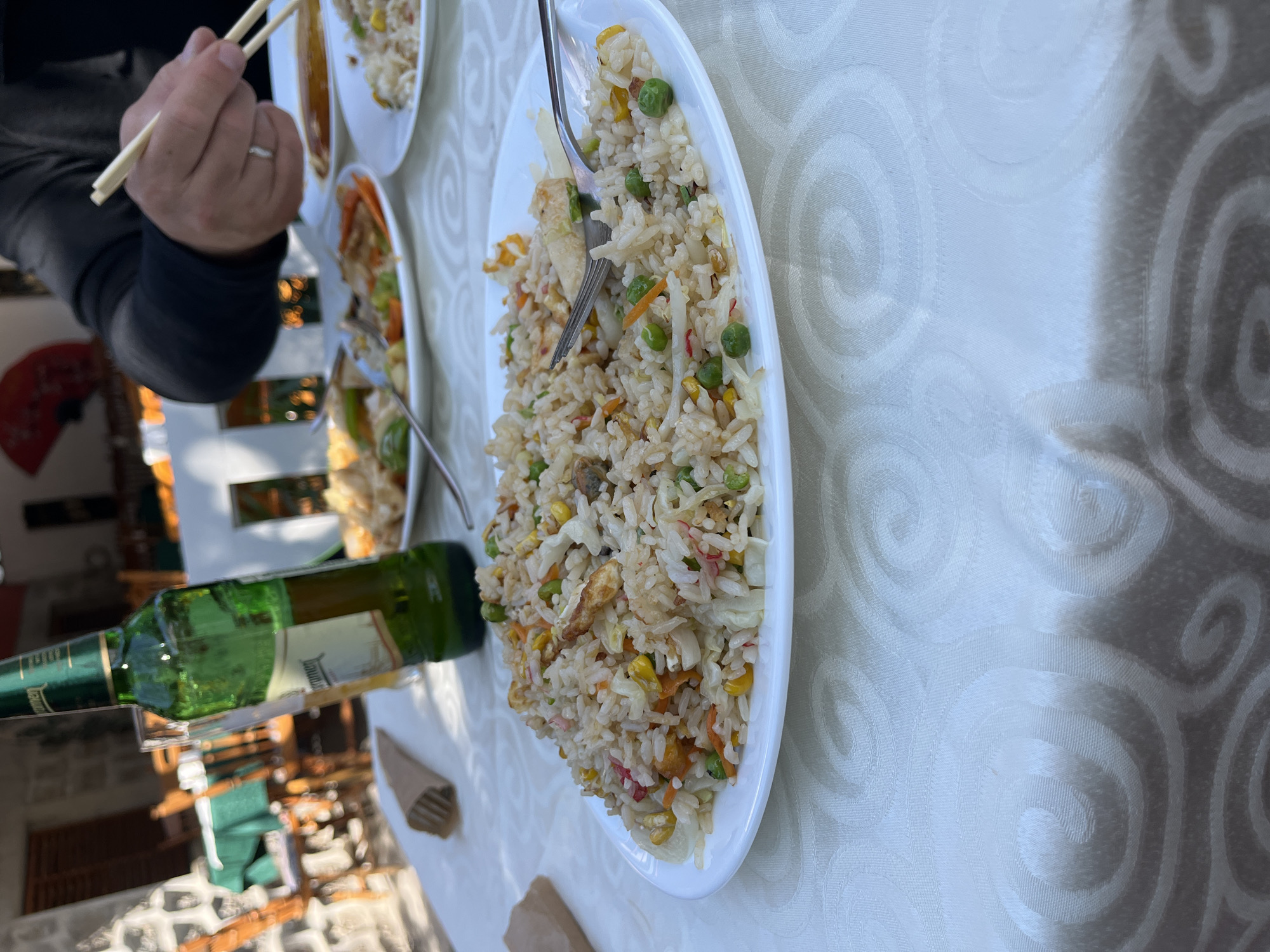 This detailed image captures an outdoor dining scene at an Asian-style restaurant in the city of Unknown, Unknown, during daylight hours.
In the foreground, a white oval plate filled with fried rice occupies the center of the frame. The rice contains visible ingredients such as green peas, yellow corn kernels, diced orange carrots, shredded white cabbage, small pieces of light-colored protein (possibly chicken or crab stick), and some thin red strands. A silver-colored fork is partially resting in the rice on this plate. To the immediate left of this plate, a folded brown paper packet, likely containing additional cutlery, lies on the white tablecloth.
The tablecloth features a subtle, embossed or woven spiral/scroll pattern.
Moving towards the mid-ground, a tall green glass bottle of Staropramen beer stands upright on the table, with a clear glass next to it. To the right, a person's right arm and hand are visible, clad in a dark, long-sleeved shirt. A silver ring is present on the ring finger. The hand is holding a pair of light-colored chopsticks, poised over a white round plate containing what appears to be another serving of fried rice. Adjacent to this plate is a small white dish holding a dark liquid, likely a dipping sauce such as soy sauce. Further back on the table, another white plate is visible, holding a stir-fried dish with various vegetables, including green bell peppers and carrots.
The background shows elements of an outdoor patio setting. Visible structures include stone wall sections and what appear to be wooden chairs with green upholstery at other tables. A prominent red, open fan is mounted as decoration on a wall. The scene is brightly illuminated by natural sunlight, casting subtle shadows and creating highlights on the objects.