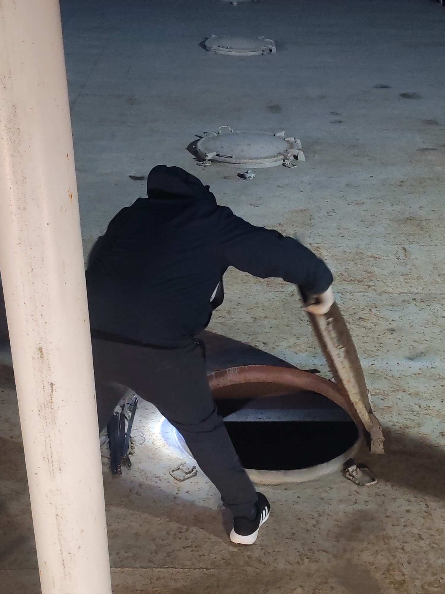 The image captures a person in a dimly lit, industrial-looking setting, likely an interior space or a covered area with a concrete or metal floor. The subject, seen from behind, is dressed in a dark hooded sweatshirt, dark trousers, and black sneakers with white soles and stripes. They are actively engaged in opening a large, circular, rusty metal hatch embedded in the floor, leaning over it with both hands on the lid. The open hatch reveals a dark, deep void beneath.
In the background, two other similar circular hatches, appearing closed, are visible on the floor, suggesting a series of access points. A prominent white pipe runs vertically along the left side of the frame, showing some dirt and wear. Near the opened hatch and the pipe, some equipment, possibly tools or cables, are scattered on the ground. A bright spot on the floor near the open hatch suggests a flashlight or other artificial light source illuminating the immediate work area. The overall lighting is artificial and somewhat subdued, indicating it might be night or a poorly lit indoor environment. The scene portrays a practical or maintenance task being carried out in what appears to be an industrial facility or a vessel in Shcheleiki, Russia. No visible text is present.