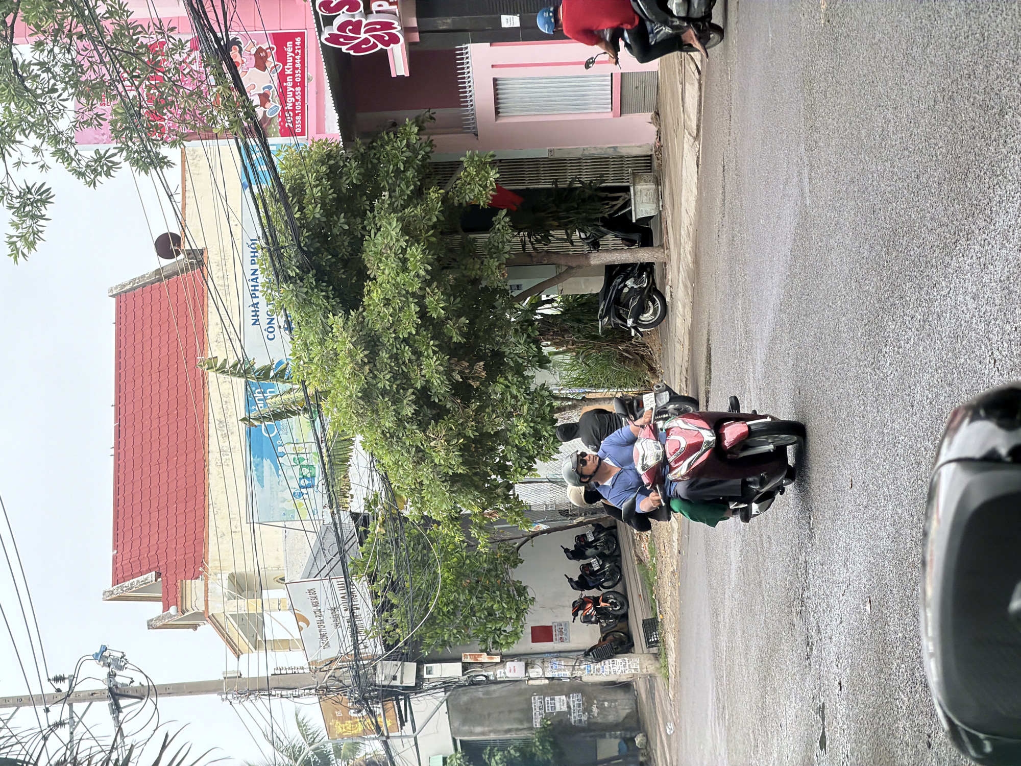 The image shows a street scene in Nha Trang, Vietnam. In the foreground, a person is riding a red scooter. The rider is wearing a blue shirt, sunglasses, and a helmet. A passenger is also on the scooter, wearing a green outfit. They are riding on a wet asphalt road, indicating recent rain.
In the background, there are several buildings. One building has a red tiled roof and cream-colored walls. Another building is painted pink and has signs with Vietnamese text and images of children. There are trees and power lines visible throughout the scene, common in urban Vietnamese environments. Several motorcycles are parked on the side of the street. The overall impression is of a busy street in a Vietnamese city, likely during the day with overcast skies.