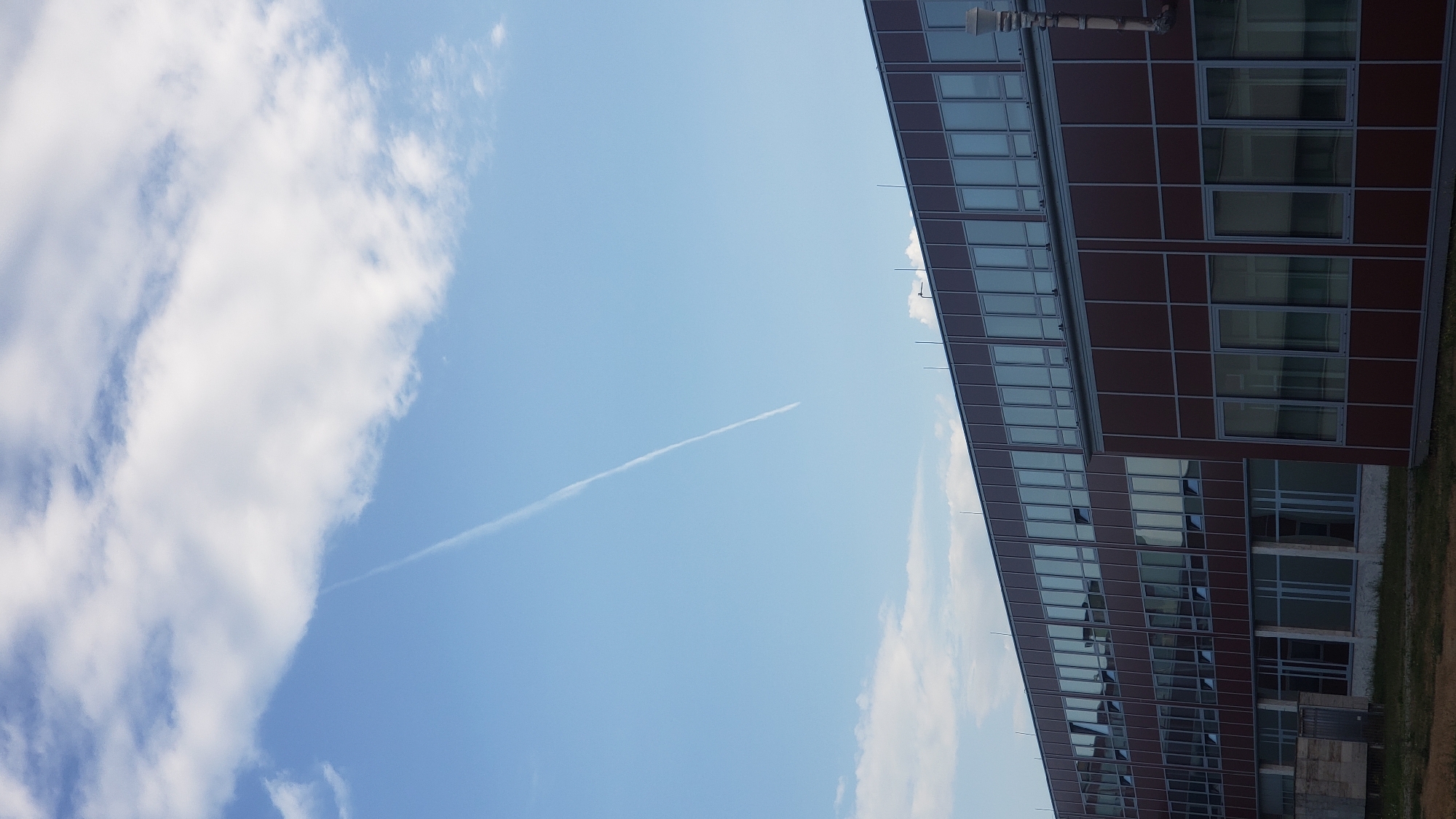 The image shows a multi-story building with a dark red facade and numerous windows under a bright blue sky with scattered white clouds. A thin contrail stretches diagonally across the sky, originating from the upper left quadrant and extending towards the lower right. The building appears to be an office or institutional structure, with a flat roof and a modern design. The foreground shows a strip of dry grass and some indistinct structures near the base of the building, suggesting an outdoor, possibly urban or suburban, setting. There are no people visible in the image. The scene is captured from a slightly low angle, emphasizing the size of the building and the expanse of the sky.