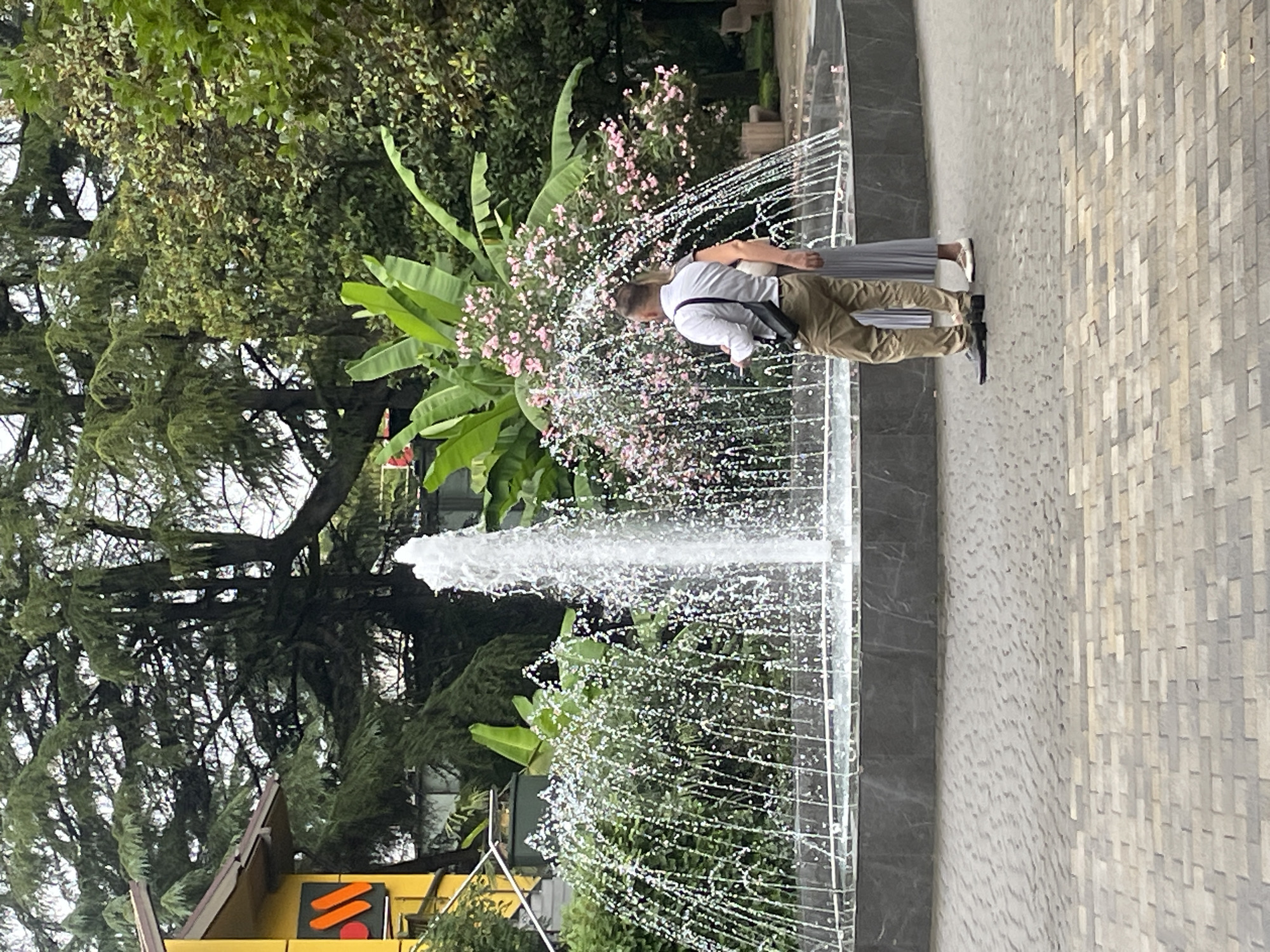 A fountain with multiple water jets is centrally located in a paved plaza. Two individuals, a man and a woman, stand near the fountain, facing away from the viewer. The man, wearing a light-colored collared shirt and khaki pants, has a black bag slung across his chest. The woman, in a light-colored pleated dress, stands beside him. In the background, lush green trees and plants, including large banana leaves and pink flowers, frame the scene. To the left, a yellow building with an orange and red logo is partially visible. The ground is paved with interlocking rectangular bricks. The overall environment suggests an outdoor public space, possibly a park or a resort area in Sochi, Russia, given the mention of the location.